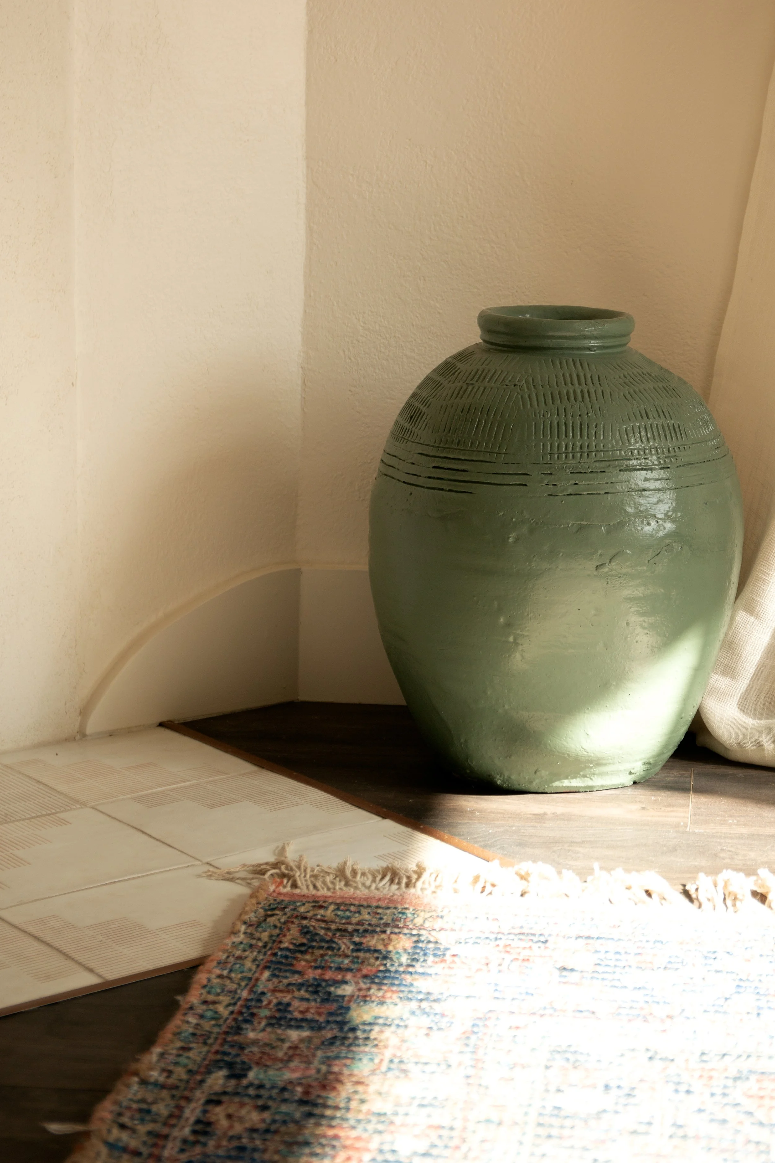 Large green ceramic vase on the floor next to a corner wall and a curtain, with sunlight casting shadows, and a patterned rug partially visible in the foreground.