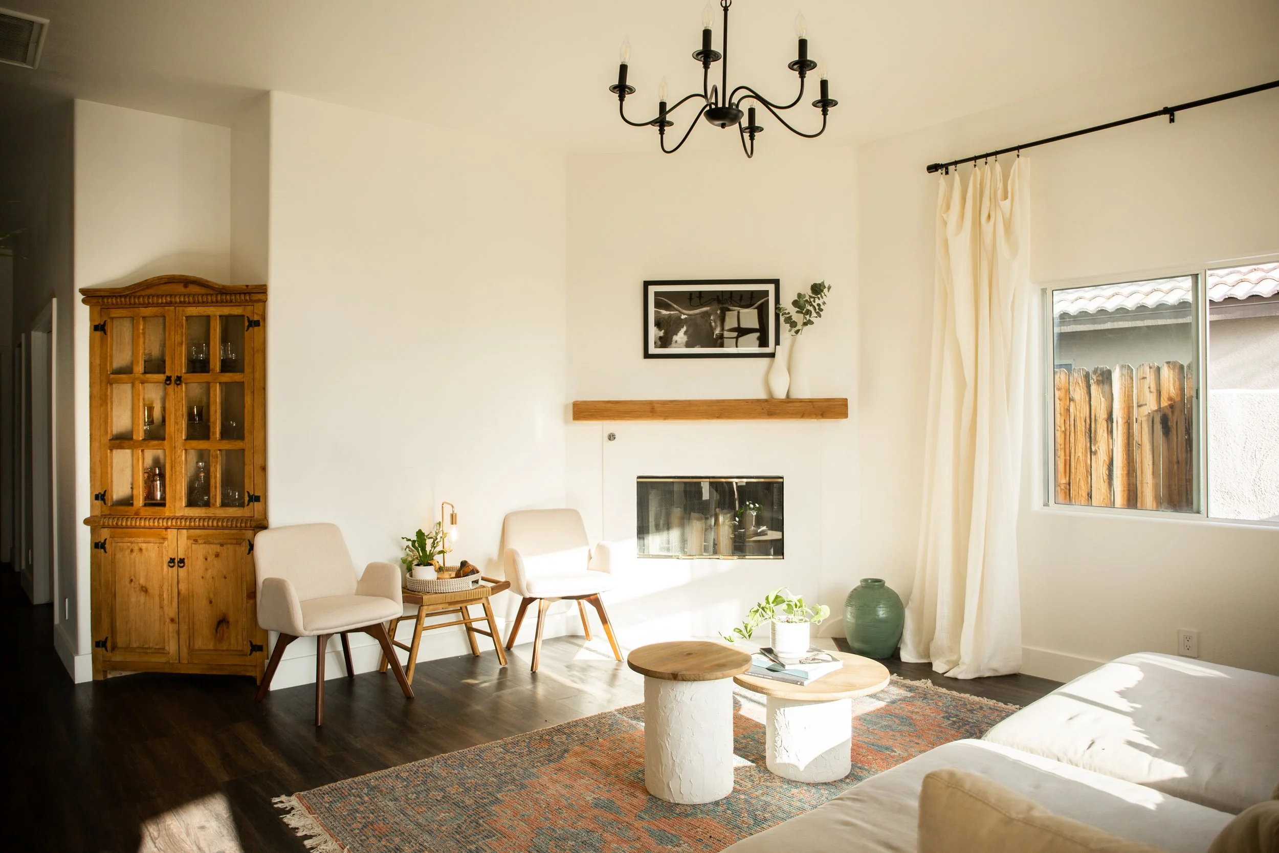 Living room with two beige armchairs, a wooden and white side table, a coffee table with a plant and magazines, a fireplace, a black and white framed photo above the fireplace, a vase with greenery, and a window with cream curtains.