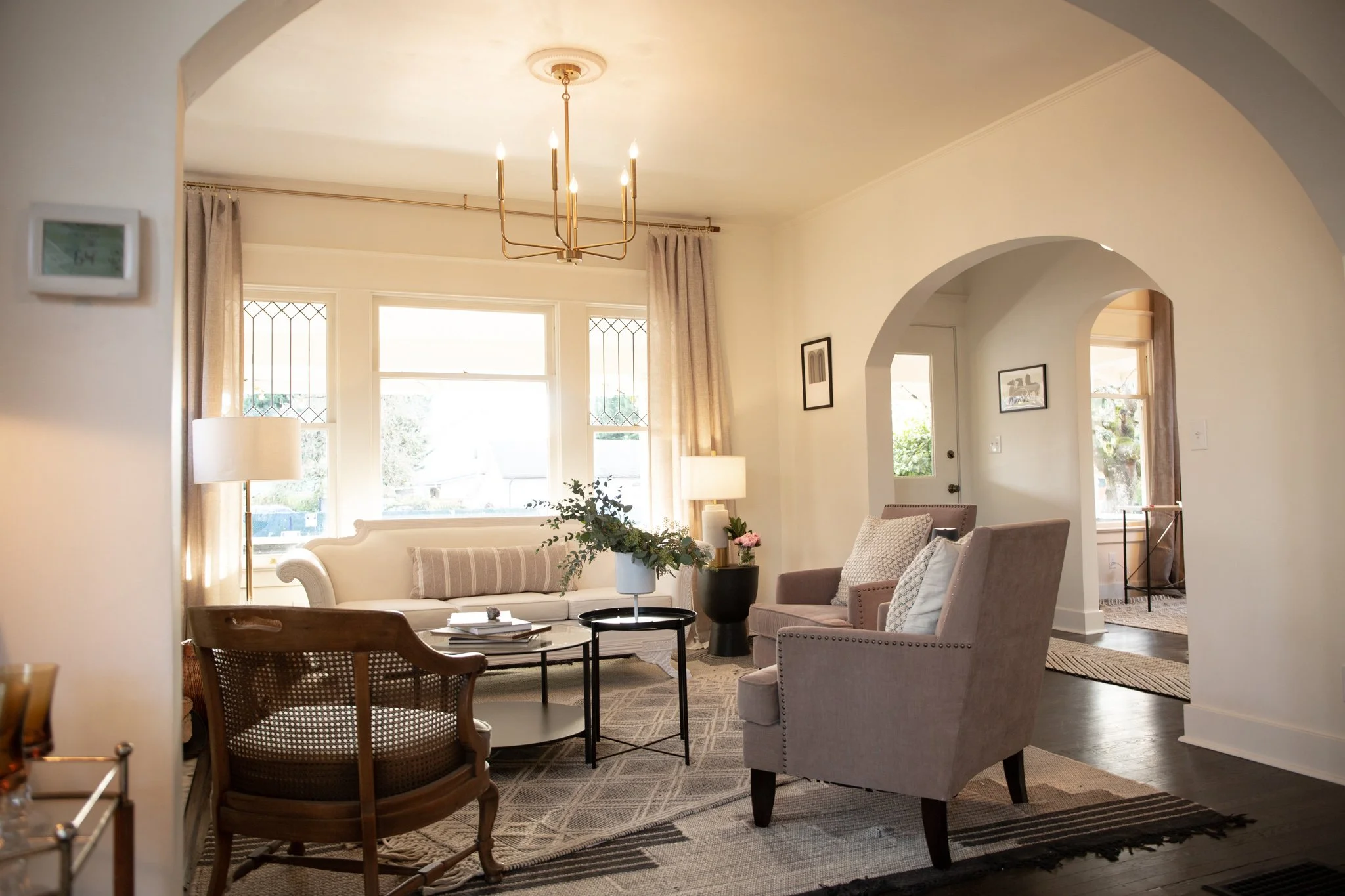 Bright living room with beige sofa, armchairs, coffee table, and floor lamps, illuminated by large windows with beige curtains, decorated with framed pictures and plants.