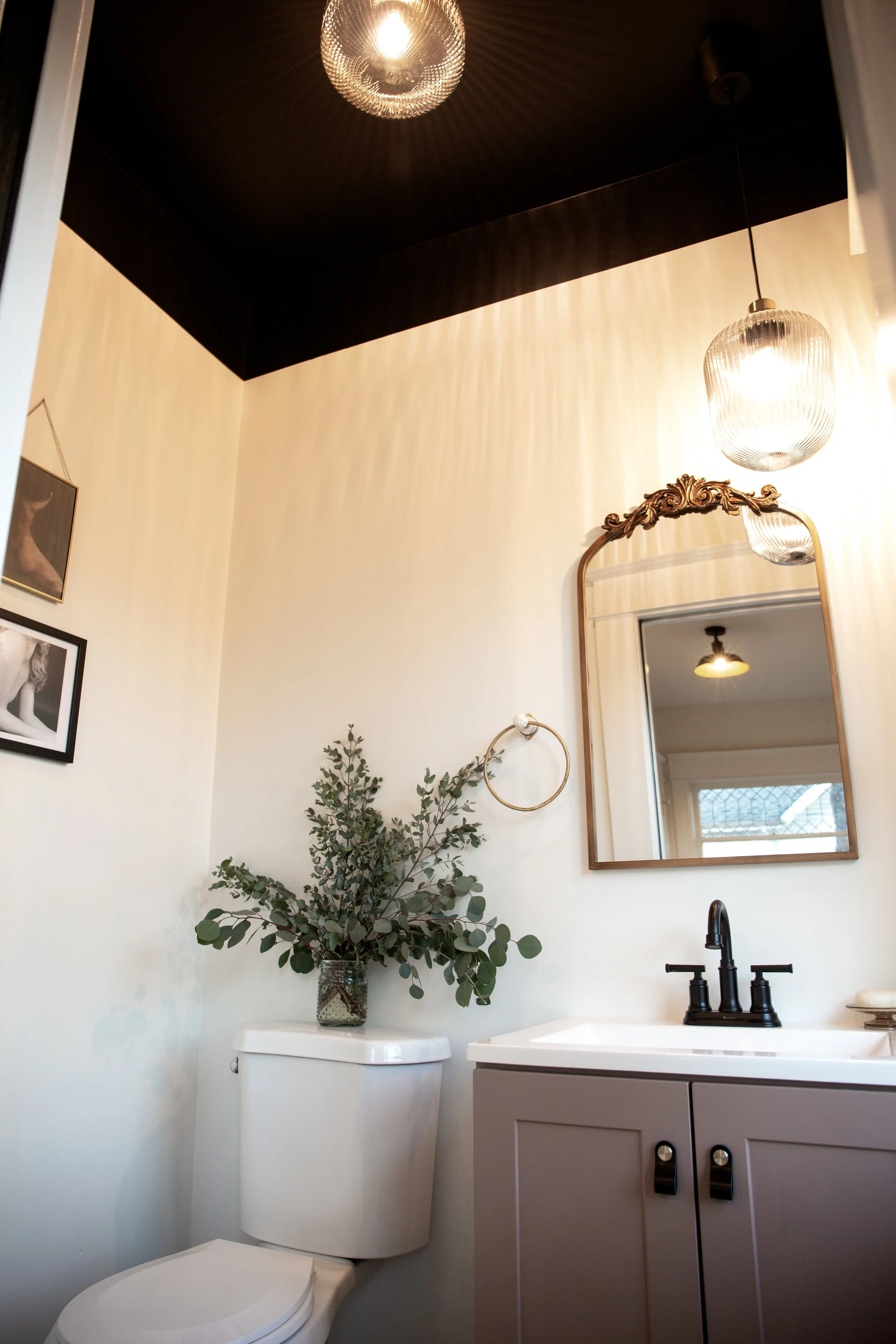 A small bathroom with a white toilet, a beige vanity with a sink, a mirror with a decorative wooden frame, two hanging light fixtures, a plant on the toilet tank, and framed photographs on the wall.