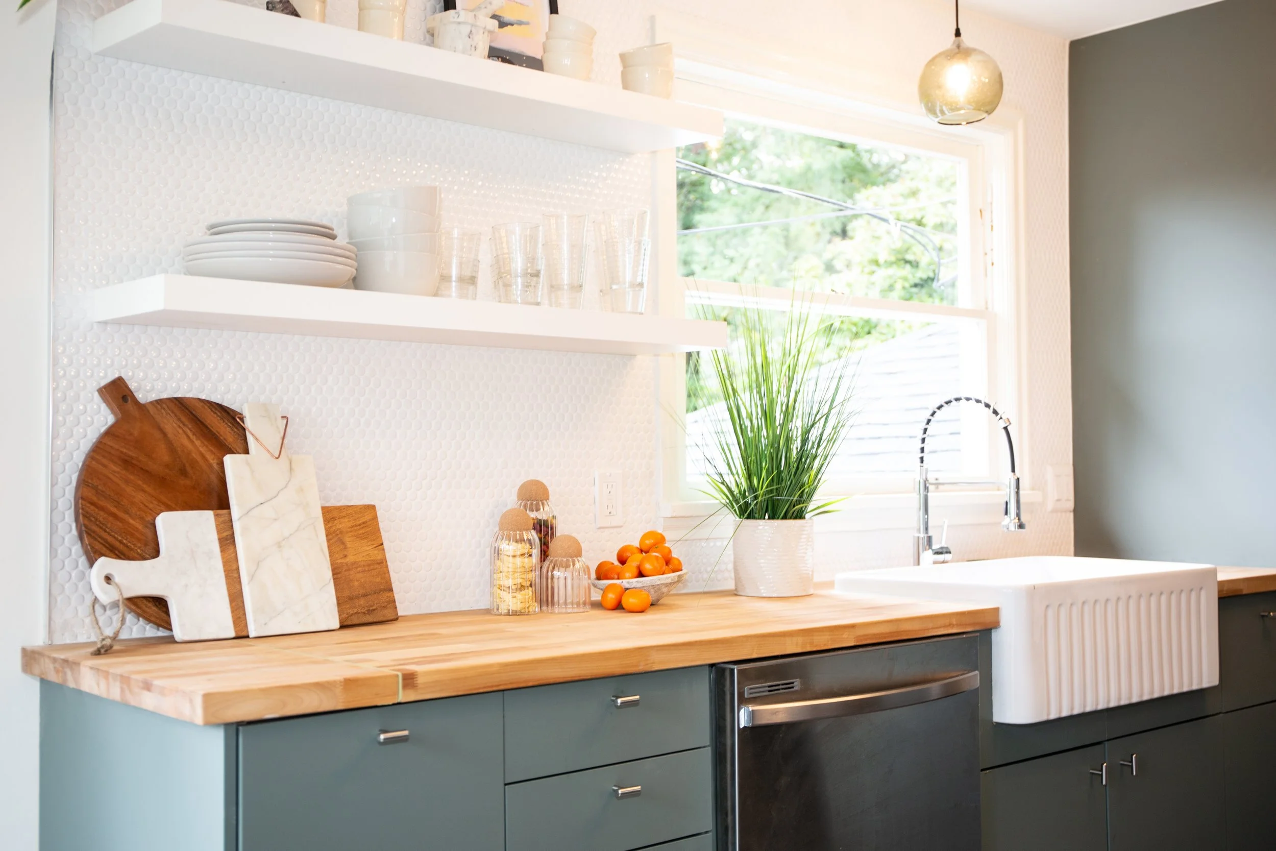 Kitchen with green cabinets, wooden countertop, white farmhouse apron sink, open window with greenery outside, and hanging pendant light. Open shelves with plates, glasses, and decorative items. Potted plant, bowls of oranges, and jars with lids on t