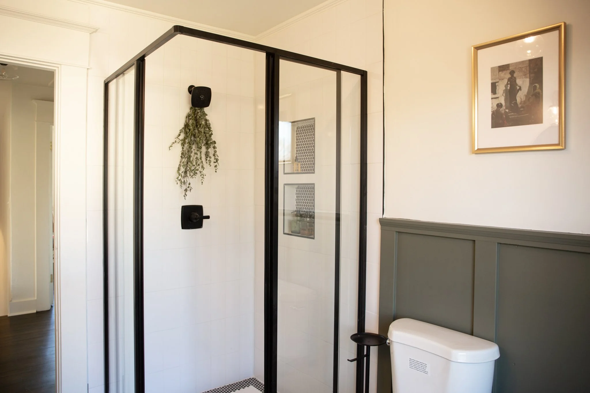 Bathroom with a glass shower enclosure, a white toilet, a framed black and white art print on the wall, and a patch of green hanging plant inside the shower.