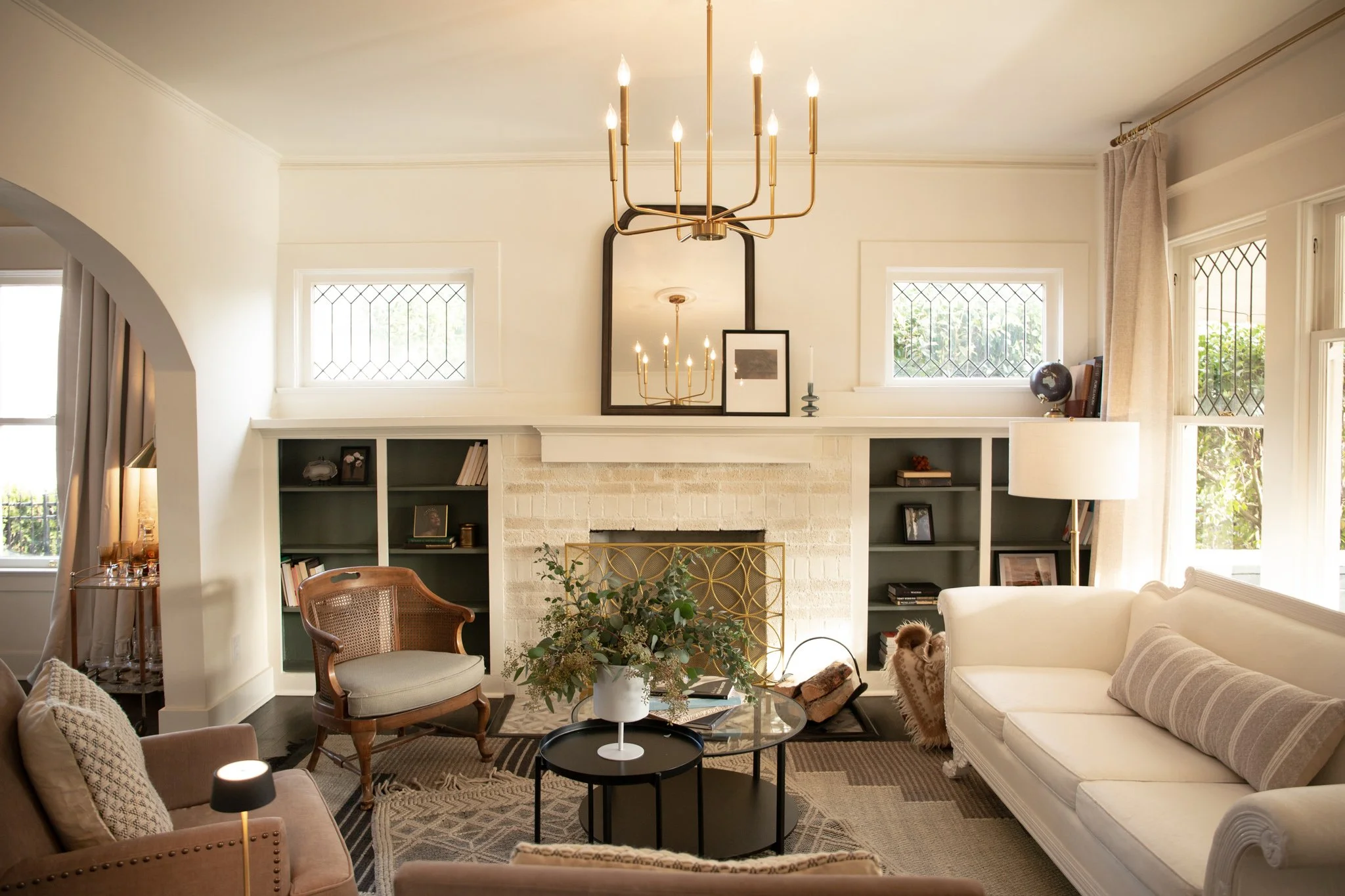 Living room with white fireplace, built-in shelves with books and decor, large mirror above fireplace, chandelier hanging from ceiling, beige sofa with striped pillow, armchair, coffee tables with decor, tall lamp, and windows with curtains.
