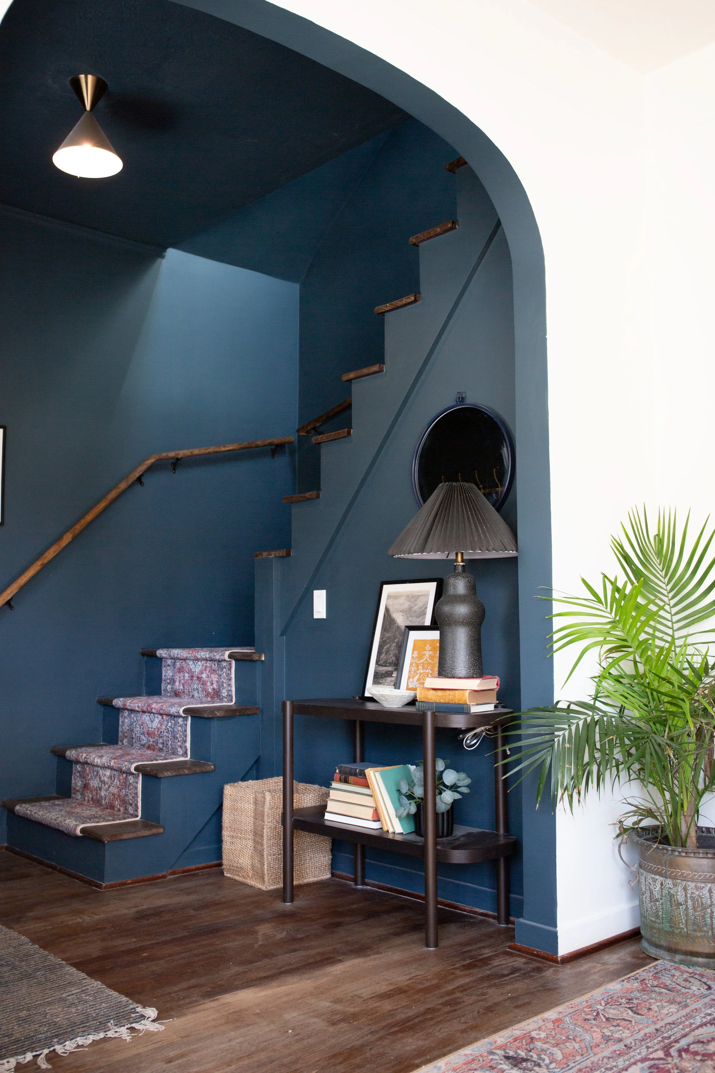 Interior of a modern home entryway with blue walls, dark wood stairs with a patterned runner, a black side table with books and framed pictures, a black table lamp, and a large potted plant.
