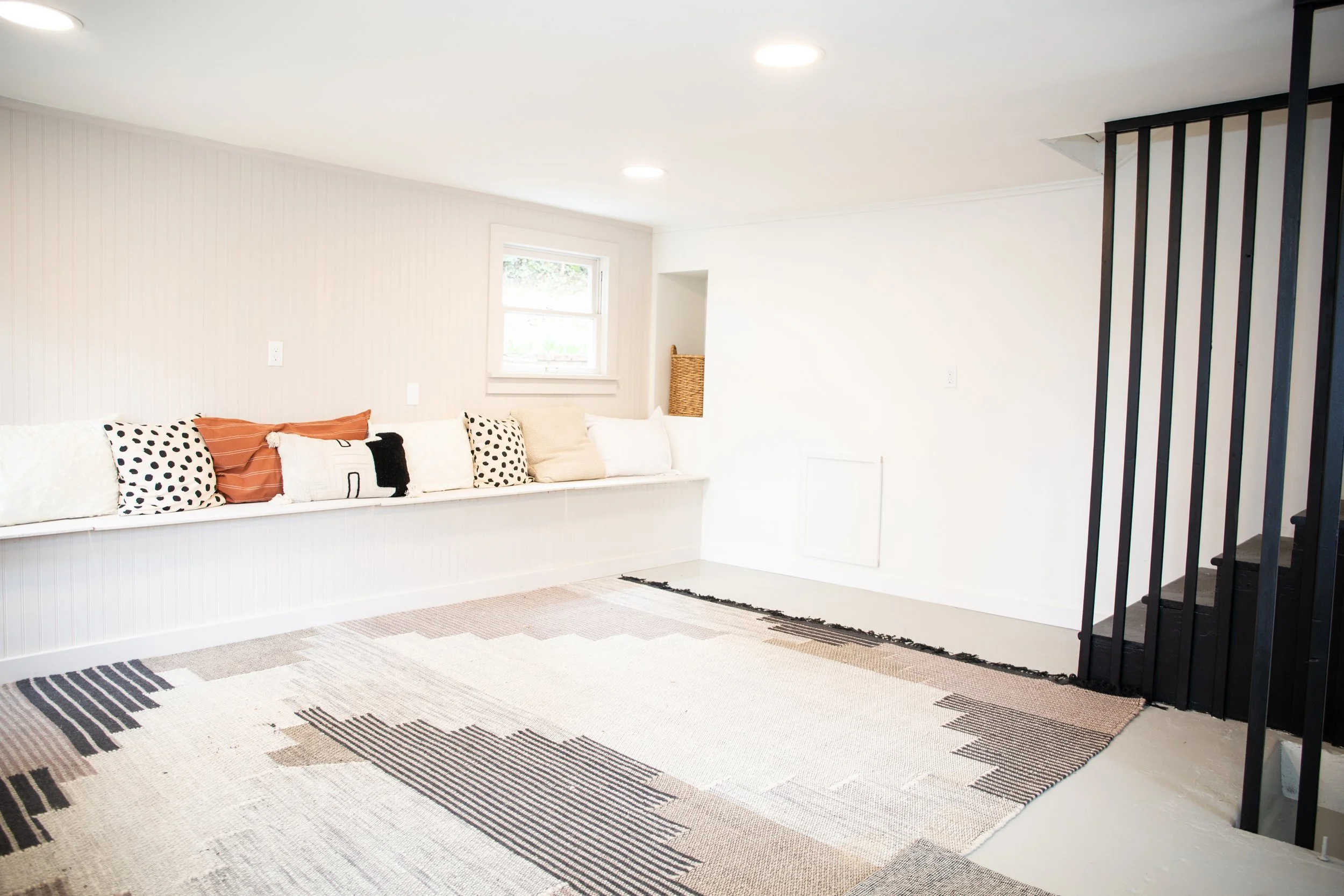Empty living room with built-in white bench with pillows, small window, beige walls, black staircase railing, and patterned area rug.