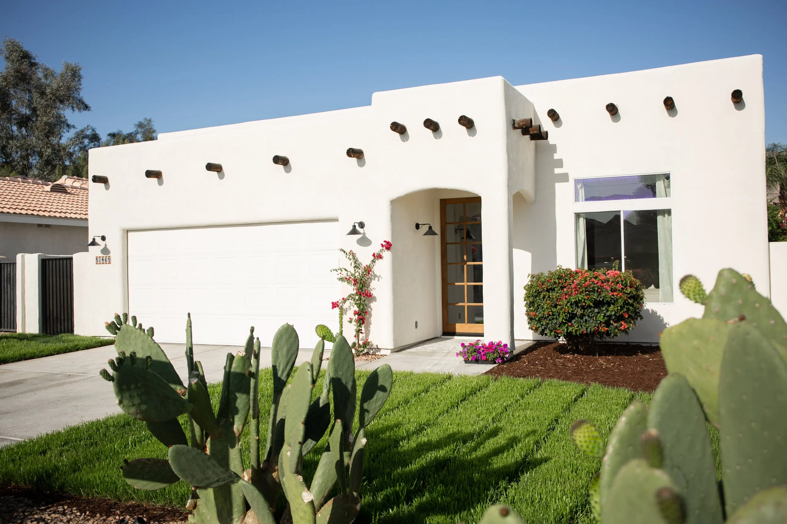 White modern house with a front yard featuring green grass, cacti, blooming bushes, and a driveway, under a clear blue sky.