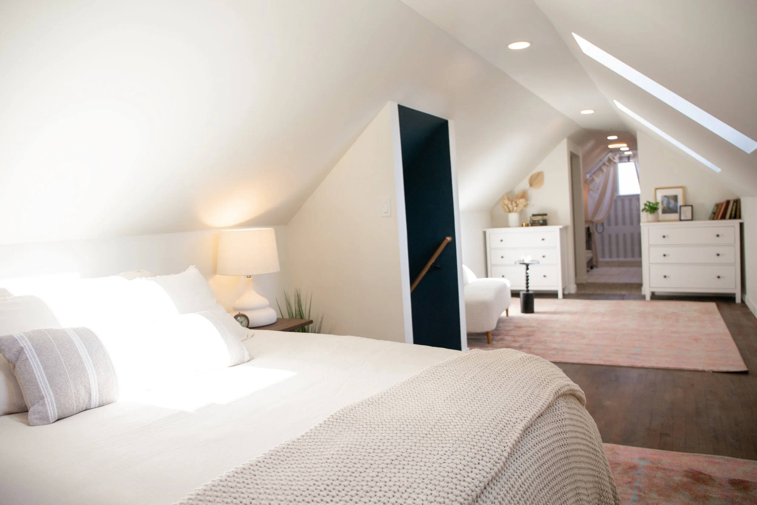 Bright bedroom with a vaulted ceiling, a bed with white bedding, a bedside lamp, and a cozy sitting area with white furniture and decor, leading to an entryway.