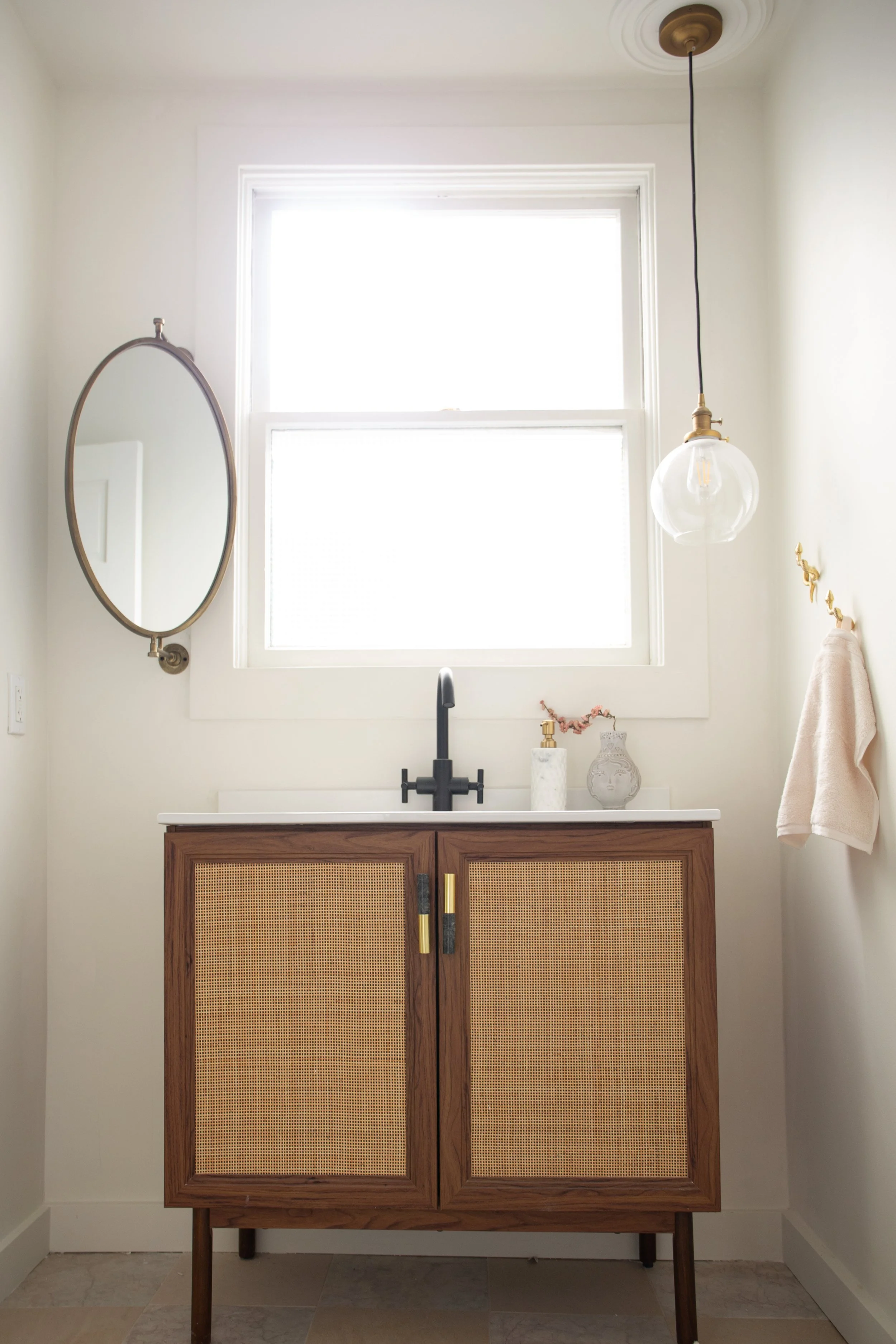 A modern bathroom vanity with a rattan door, black faucet, and decorative items under a large window, with a wall mirror and hanging pendant light.
