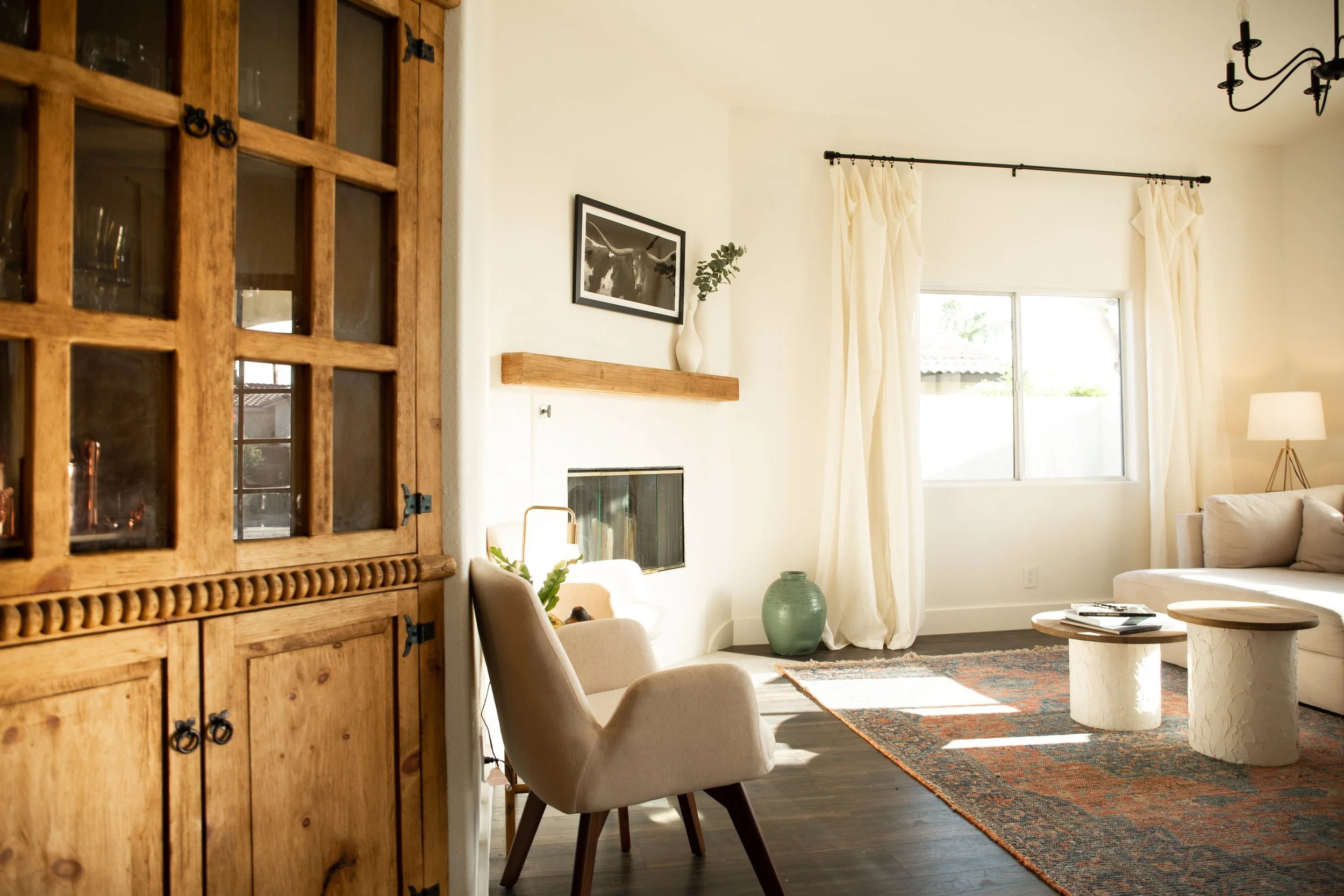 Bright living room with white walls, large window with cream curtains, cream sofa, beige armchair, rustic wooden cabinet, modern coffee tables, area rug, and decorative elements like a framed black-and-white photo, a ceramic vase, and a floor lamp.