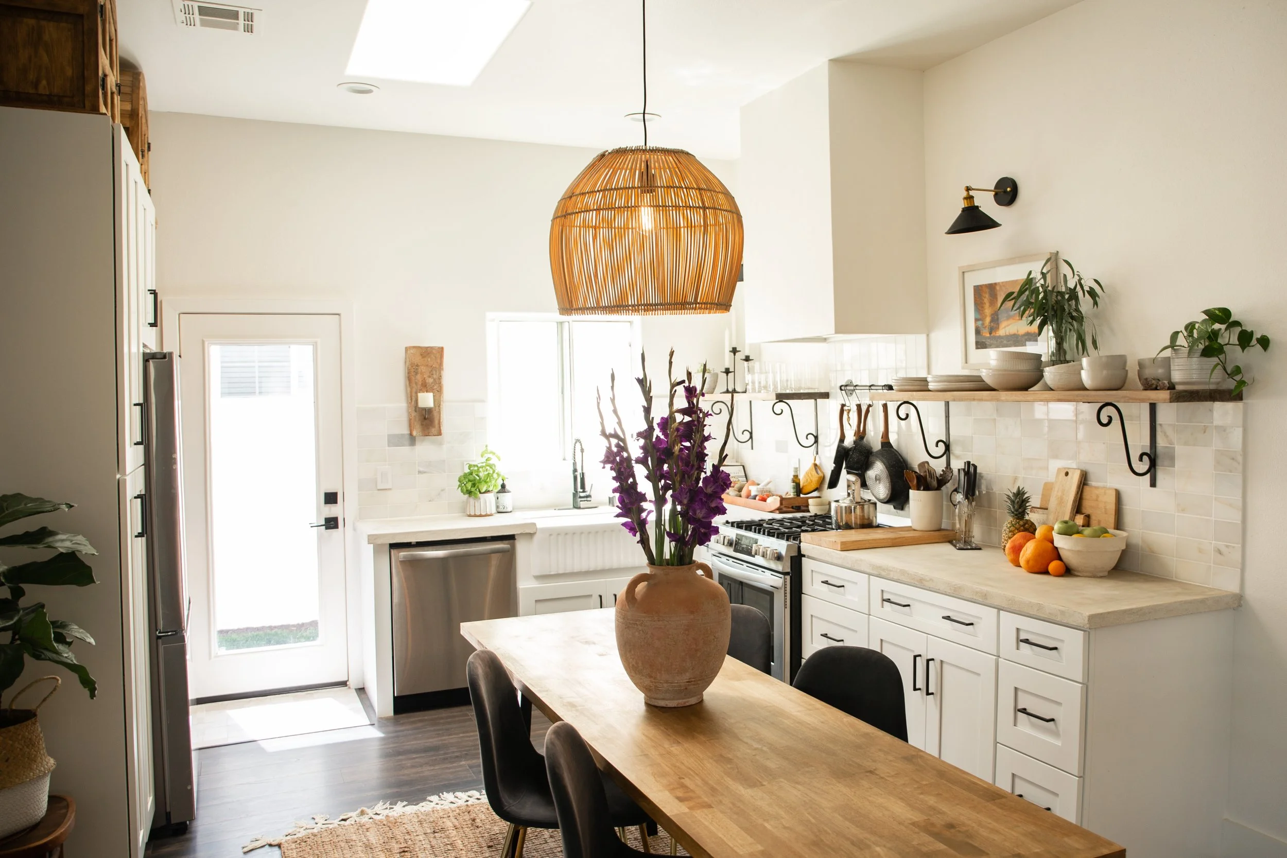 Bright, modern kitchen with white cabinets, a wooden dining table with black chairs, and a large wicker pendant light. Decor includes a clay vase with purple flowers, bowls, fruit, and potted plants.