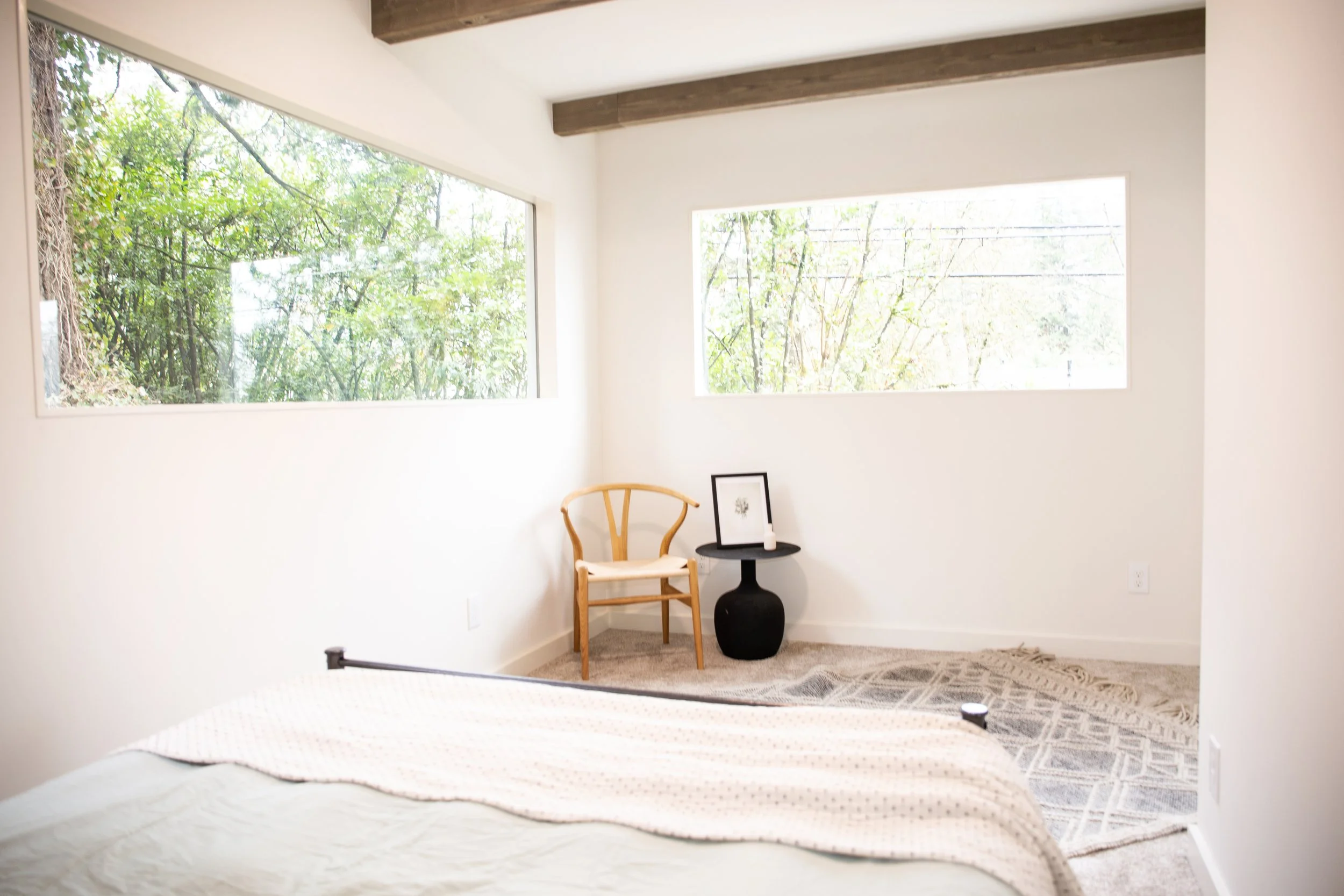 Minimalist bedroom corner with large windows showing greenery outside, a wooden chair, a small black side table with framed artwork and a candle, and a patterned rug on light-colored carpet.
