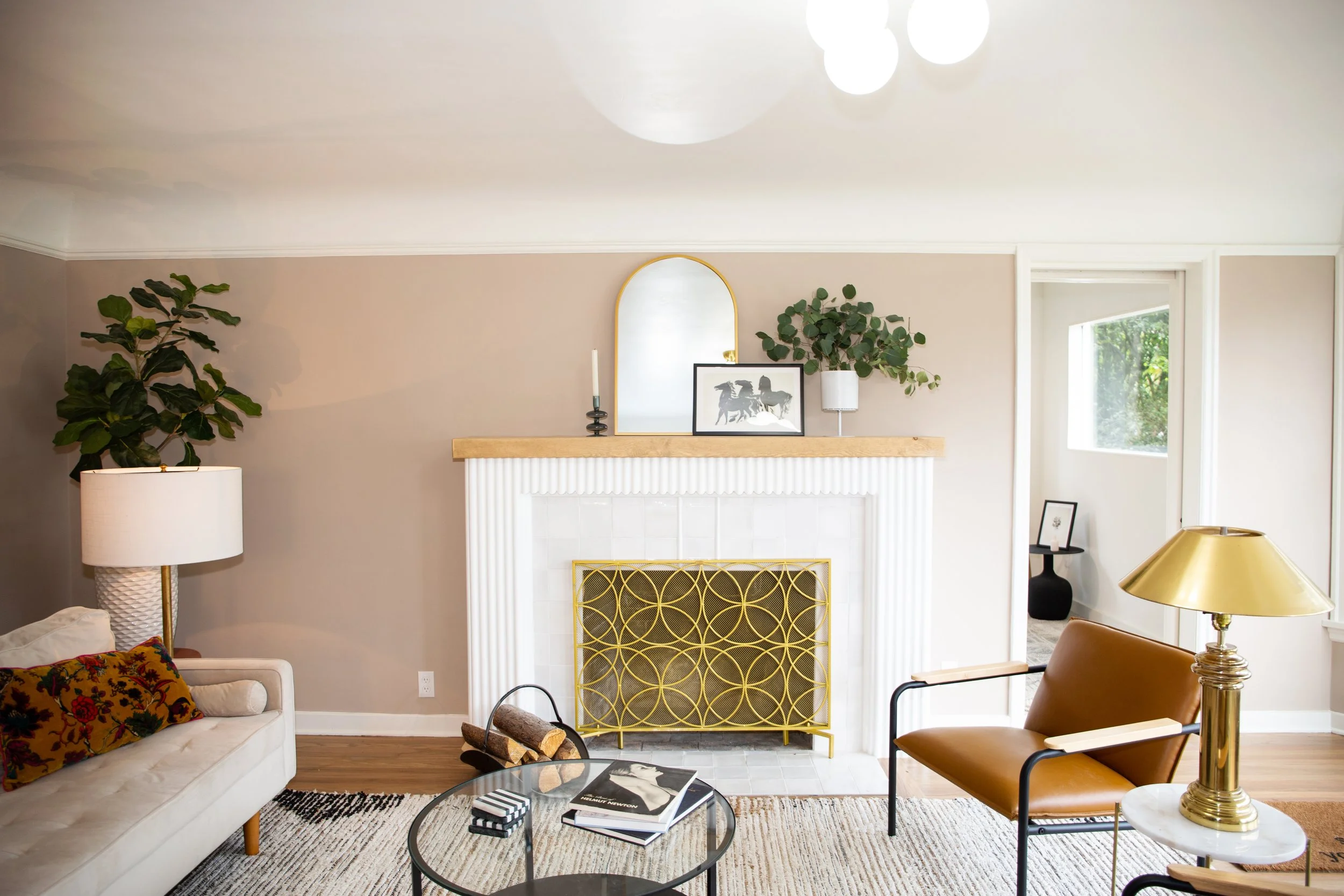 Living room with beige walls, white fireplace with gold decorative screen, wooden mantle with mirror, framed photo, candle, plants, leather armchair, patterned pillow, side table, coffee table with books and logs, large window, and various lamps.