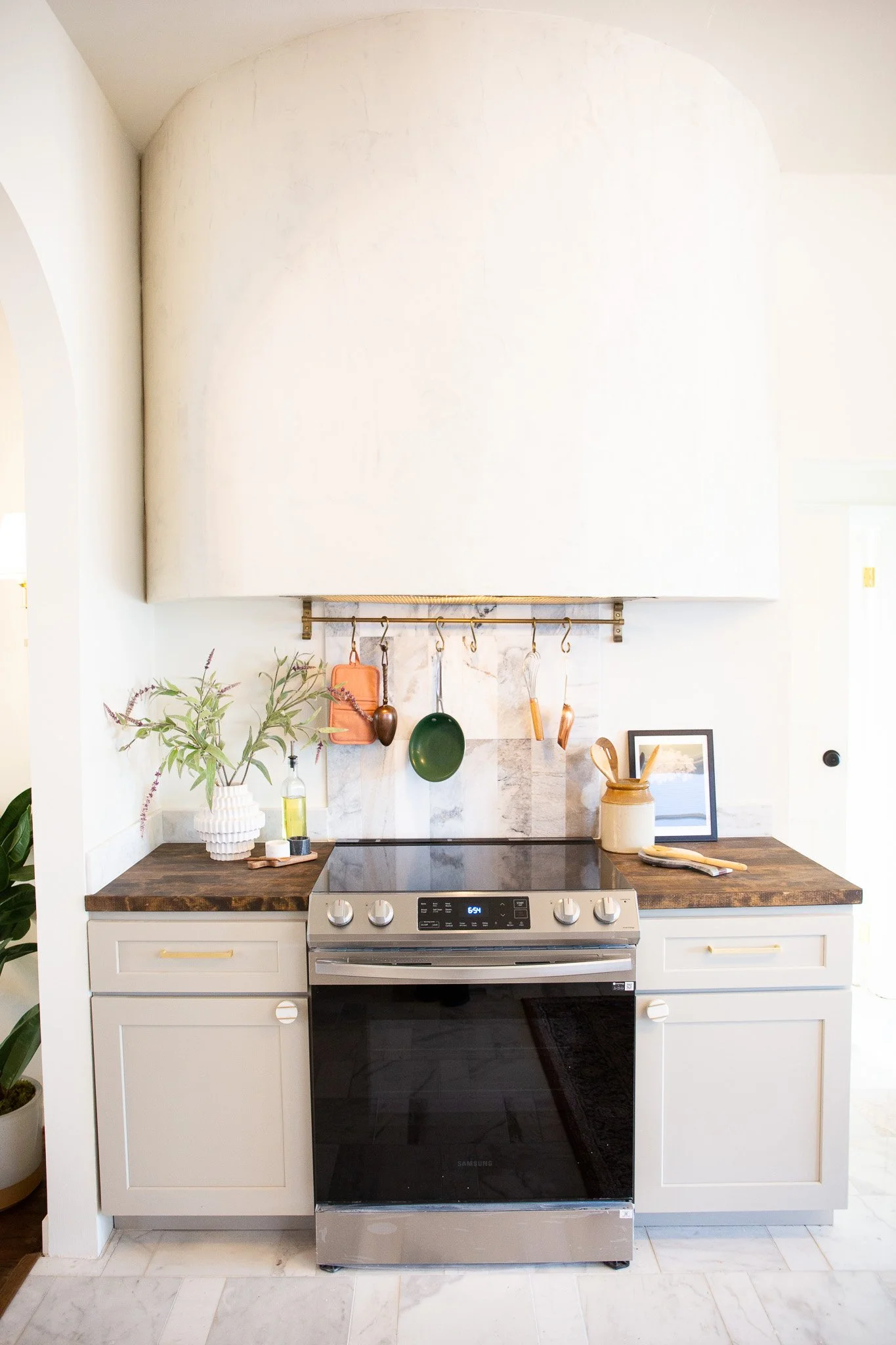 A modern kitchen with a white stove, dark wood countertops, potted plant, framed picture, and various hanging kitchen utensils.