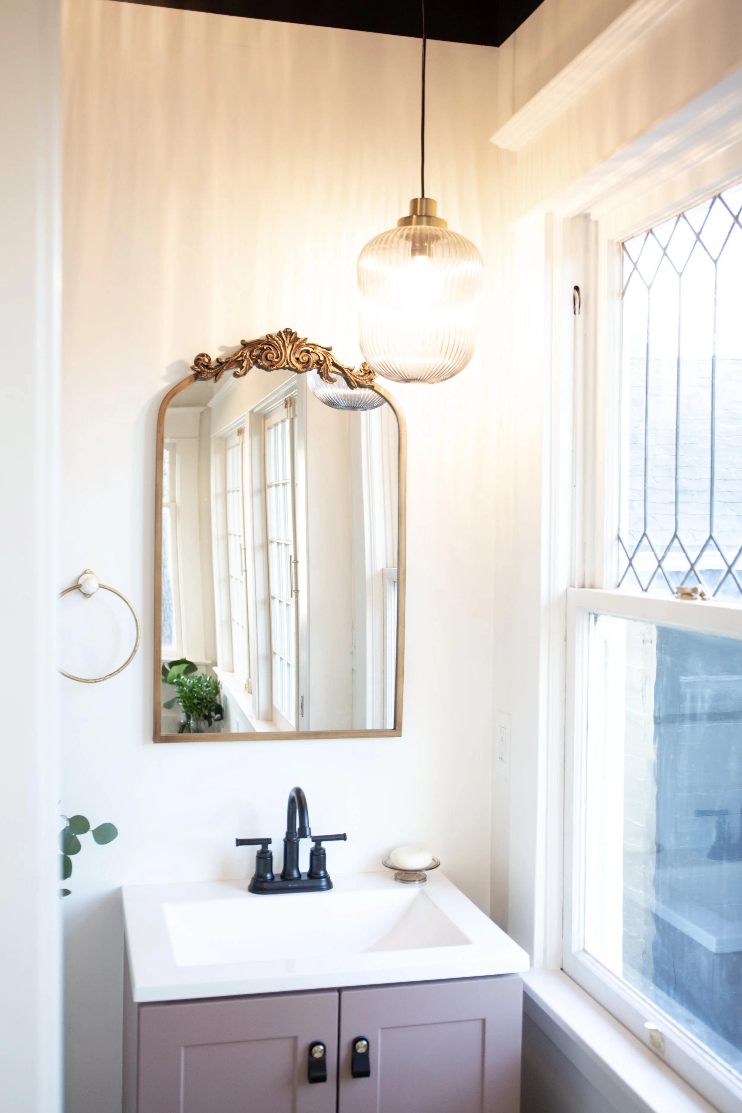 A bathroom with a white vanity, black faucet, mirror with ornate gold frame, hanging light fixture, window with diamond-shaped grid, and a potted plant on the windowsill.