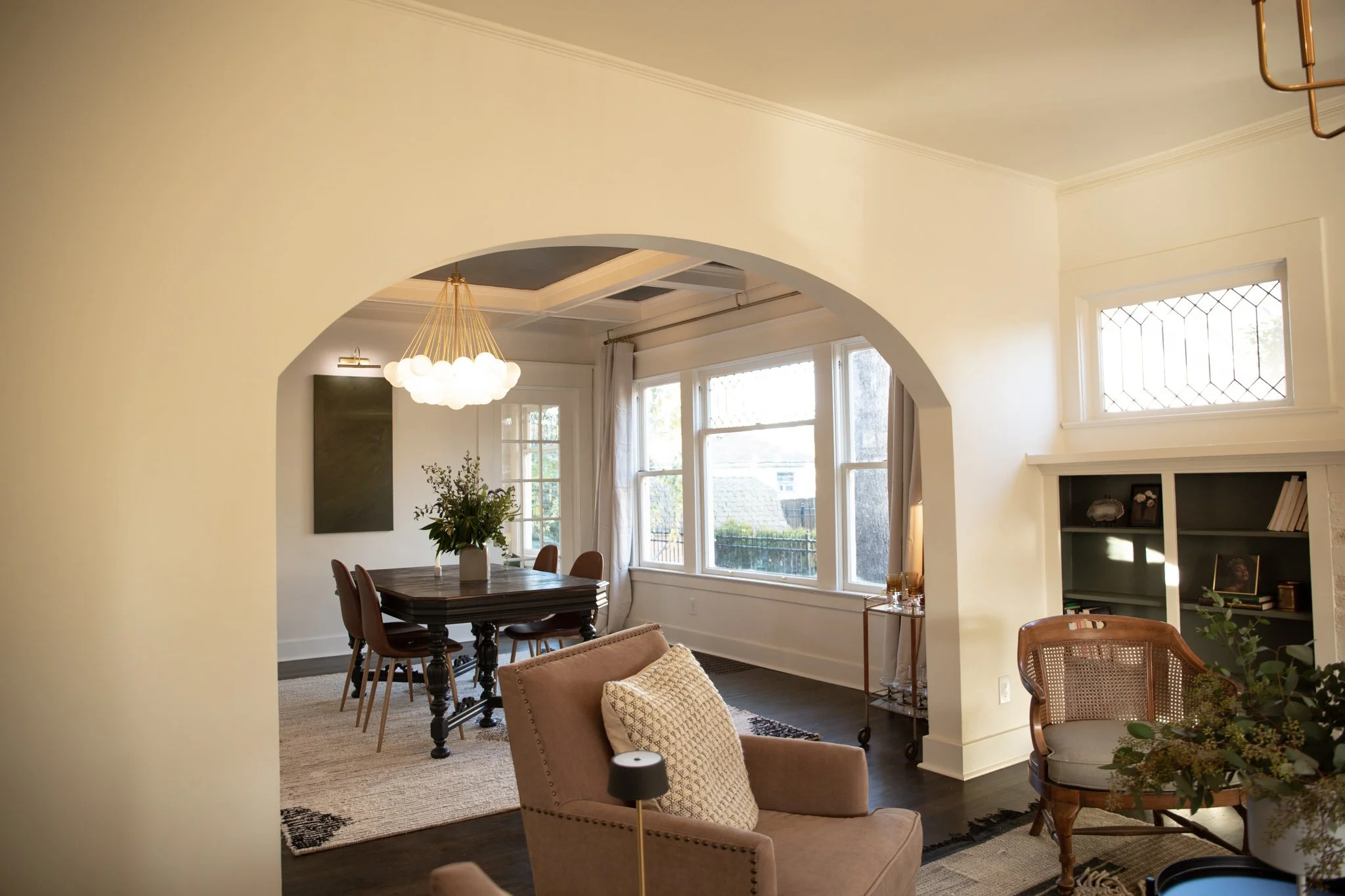 Living room and dining area with large windows, beige walls, and dark wood furniture, including armchairs, a dining table, and a bookshelf.