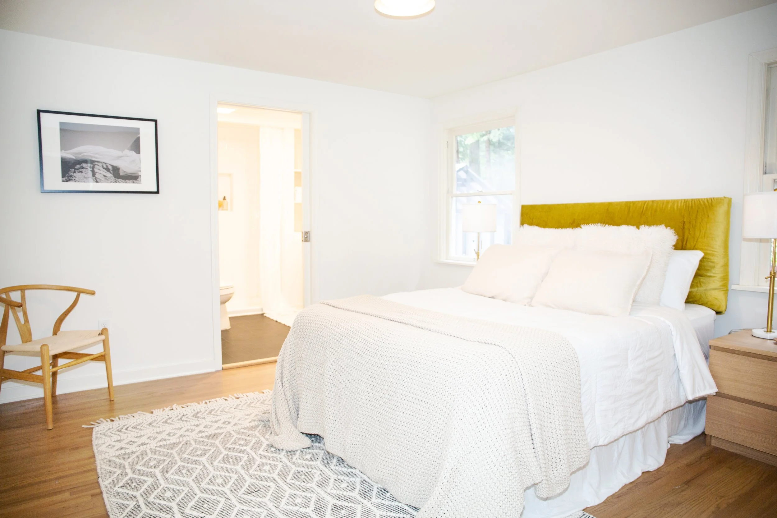 Bright bedroom with white bedding, yellow headboard, wooden nightstand, and a rug, with a visible bathroom in the background.