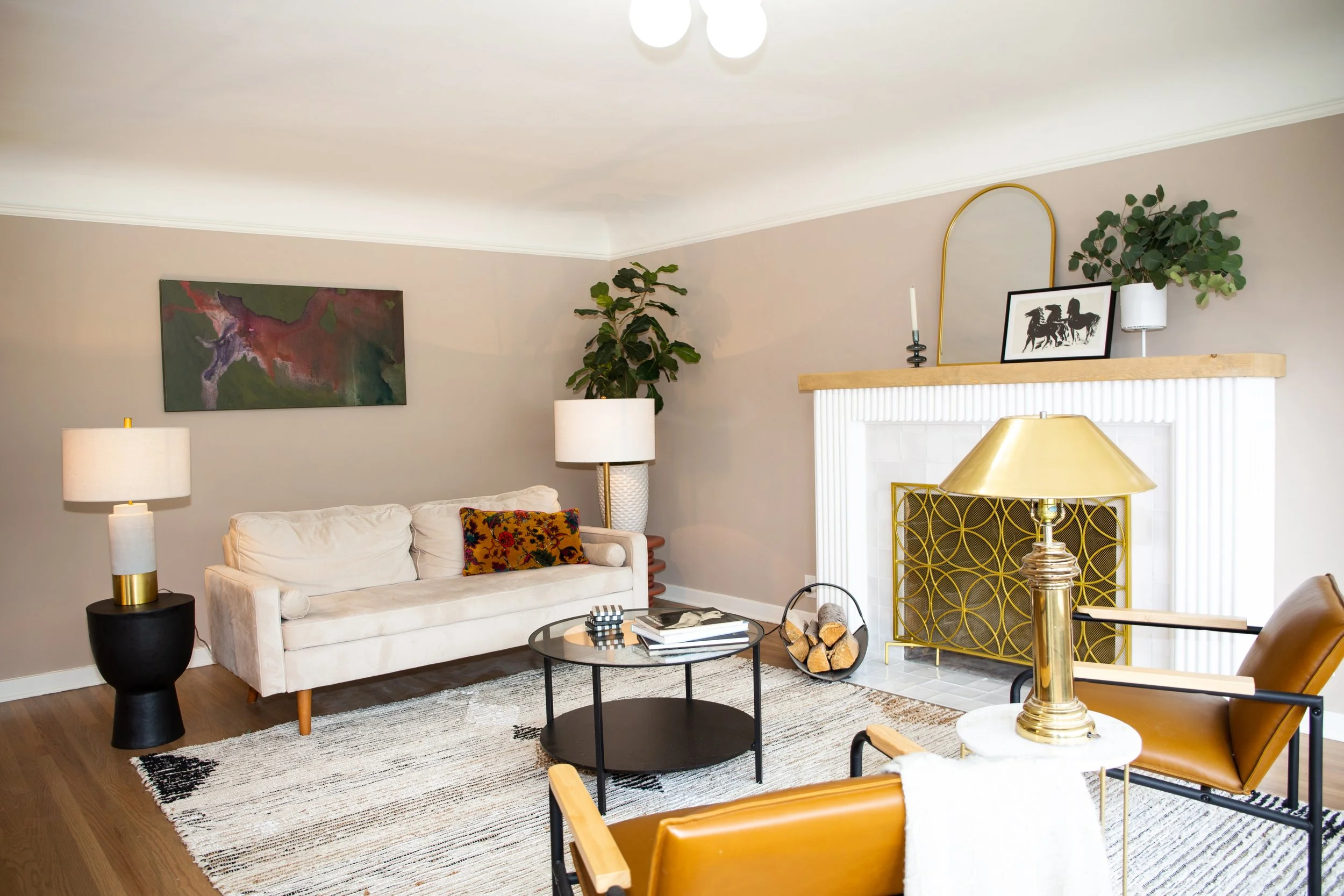 Living room with beige walls, white couch, black coffee table, tan leather chairs, modern lamps, abstract wall art, plants, fireplace with gold screen, and decorative items.