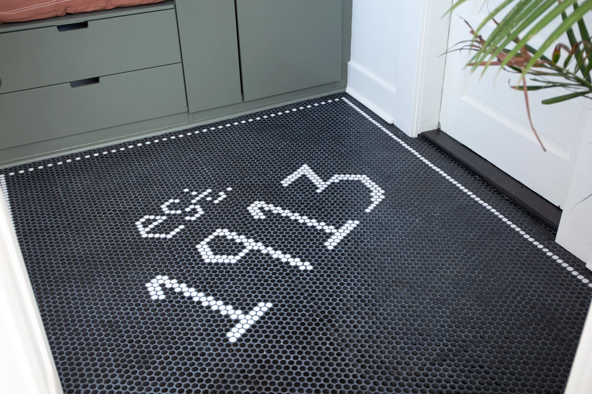 Black entryway mat with white hopscotch and dashed border near a white door and green cabinet.