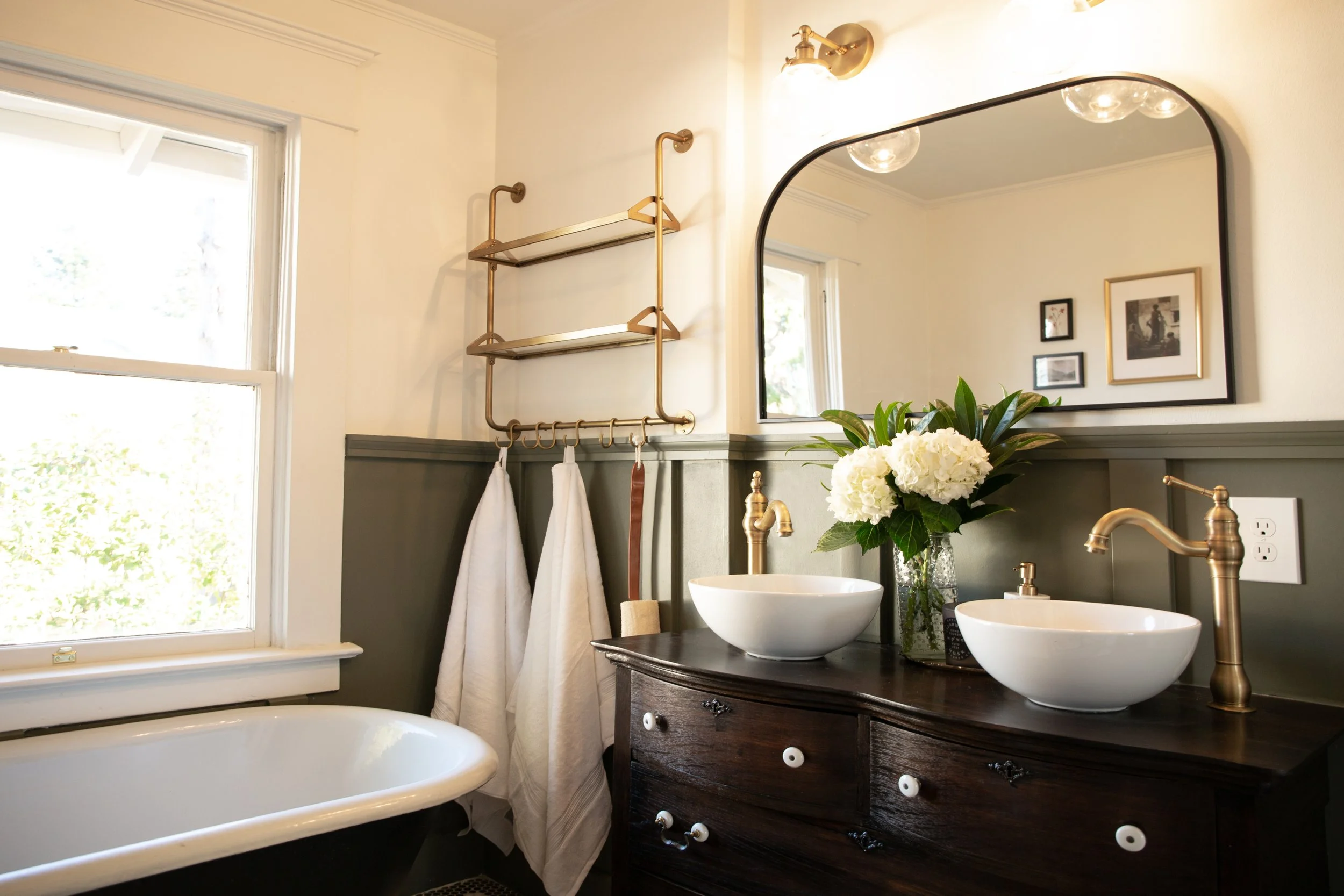 Bathroom with a window, a dark wooden vanity with two vessel sinks and gold fixtures, a large mirror, white towels hanging on a gold towel rack, a flower vase with white flowers, and framed pictures on the wall.
