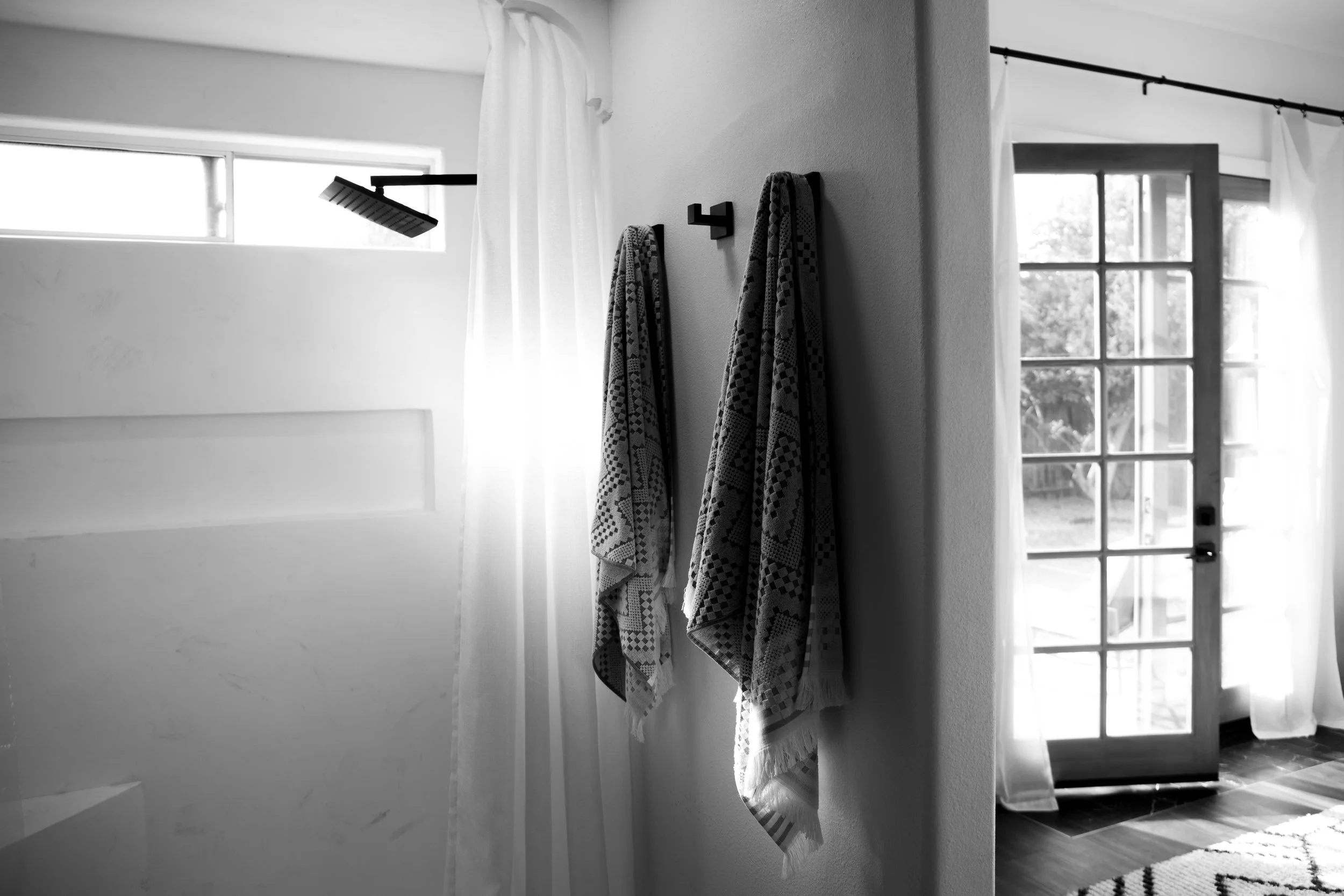 Black and white photo of two patterned towels hanging on hooks on a wall, with a bright window and a door with a glass panel in the background.