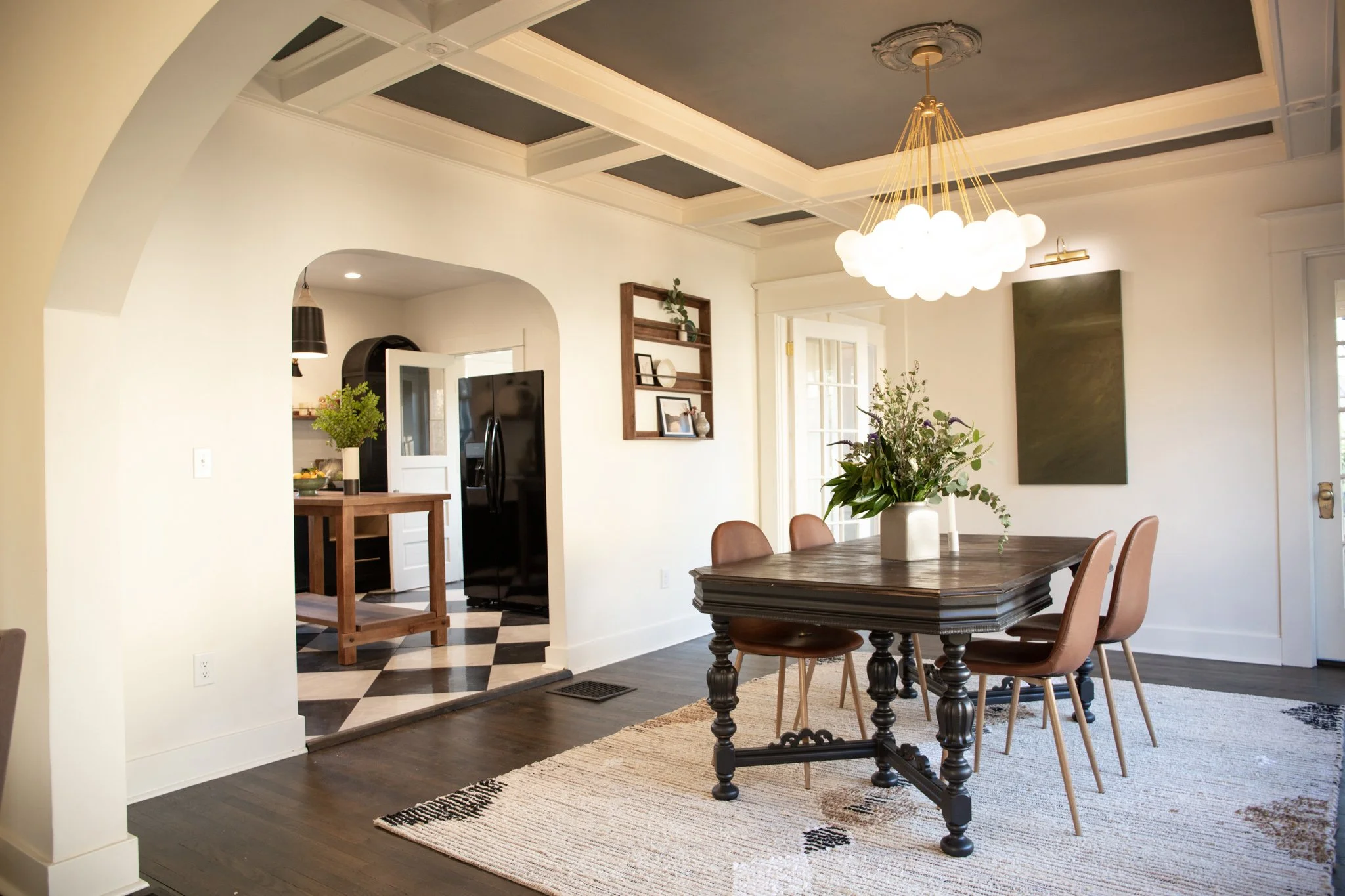 A dining room with a dark wooden table, surrounded by five brown chairs, a large white vase with green plants as a centerpiece, a white wall with a small wooden shelf, and a modern chandelier hanging from the ceiling. An art piece is on the wall, and