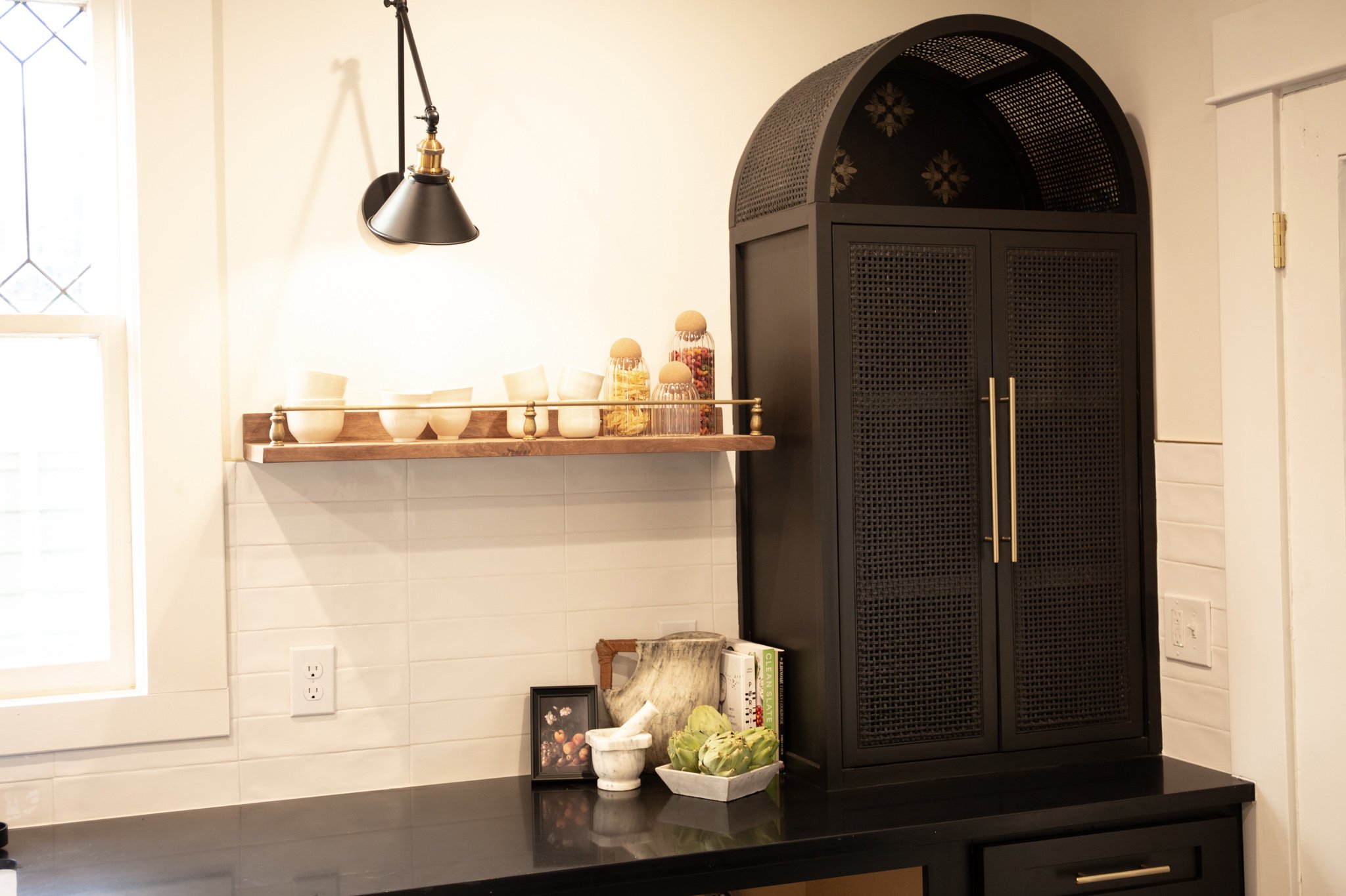Kitchen countertop with a black cabinet, a wooden shelf with bowls and jars, and decorative items, near a window with natural light.