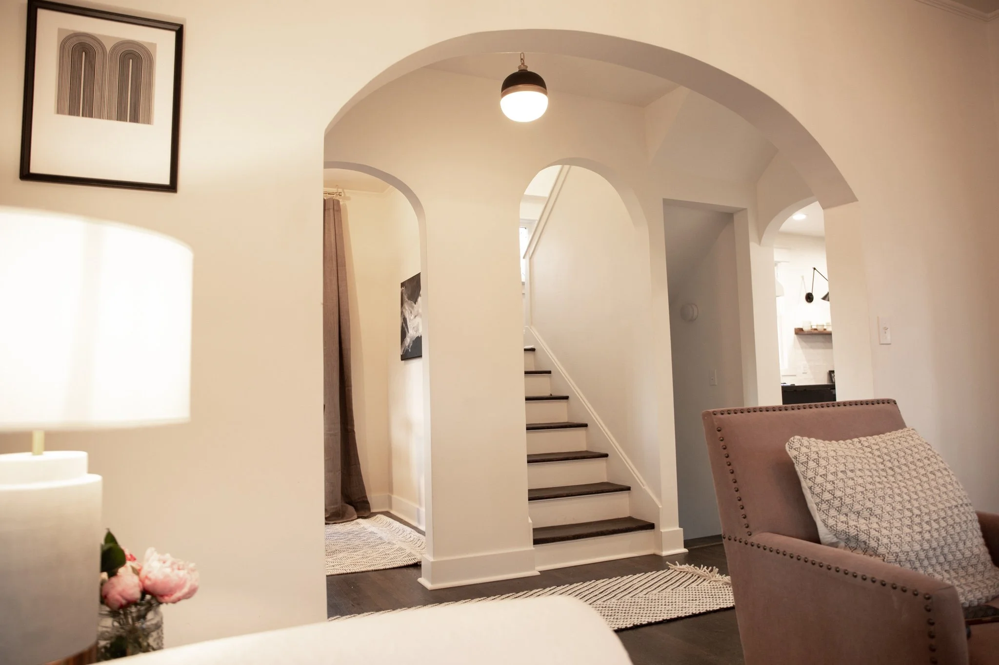 Living room with a staircase leading to the upper floor, framed artwork on the wall, and a beige armchair with a patterned throw pillow.