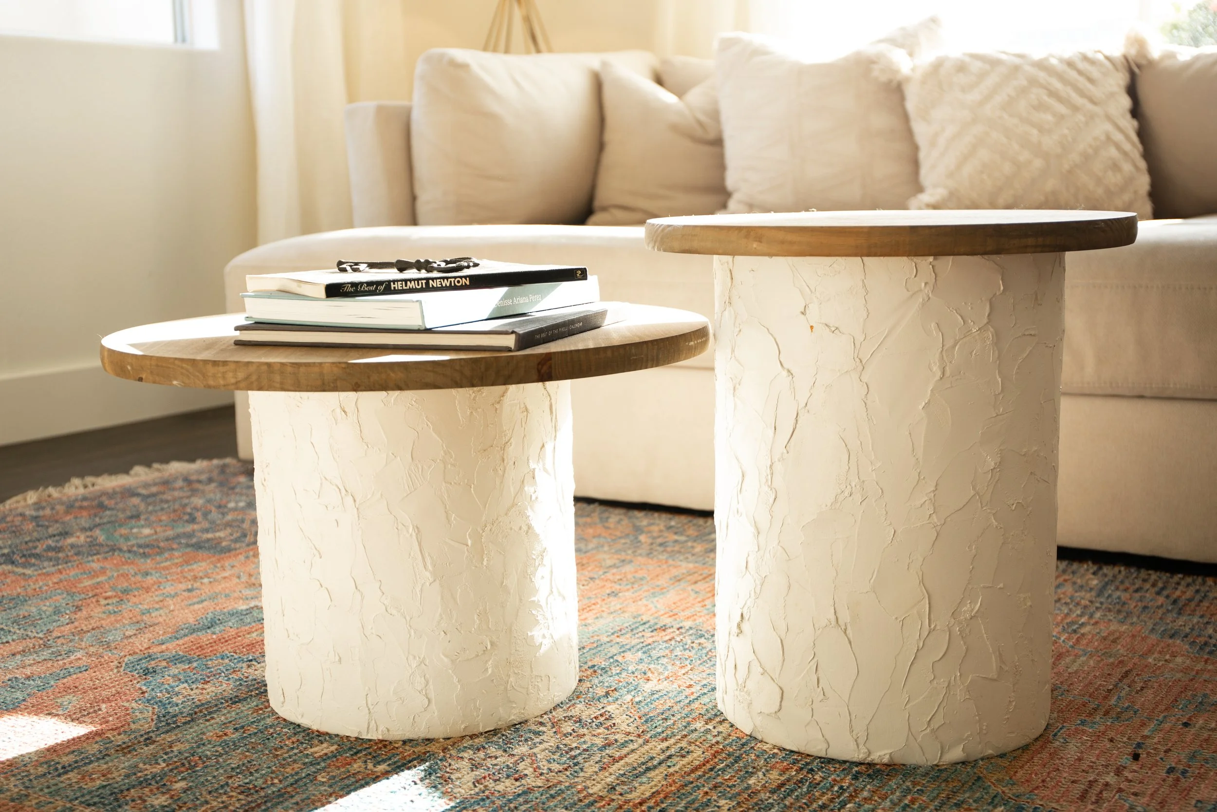 Two modern round coffee tables with textured white bases and wooden tops in a cozy living room, with a beige sofa and colorful rug in the background.