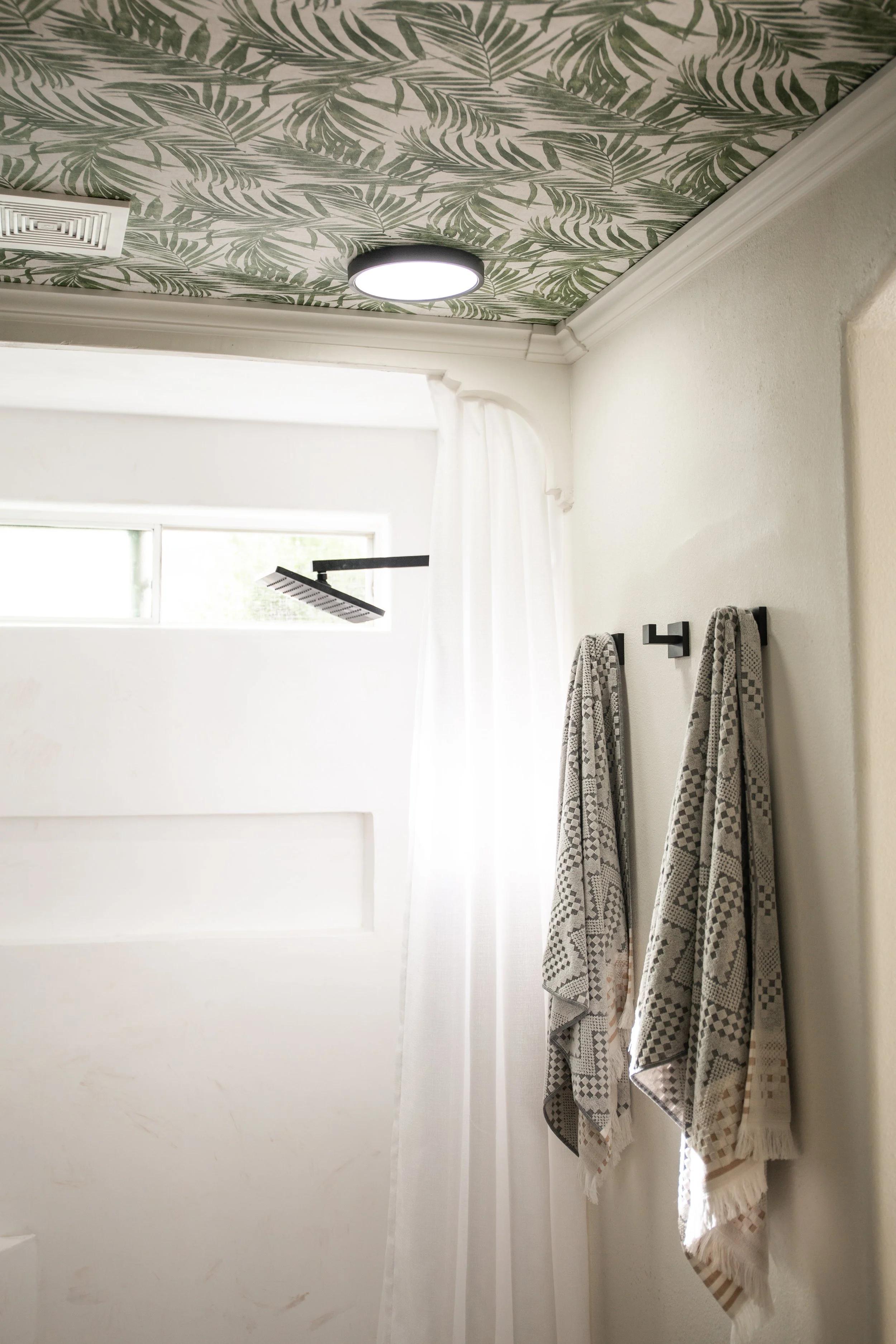 Bathroom with patterned ceiling, black towel hooks with towels, window, and showerhead.