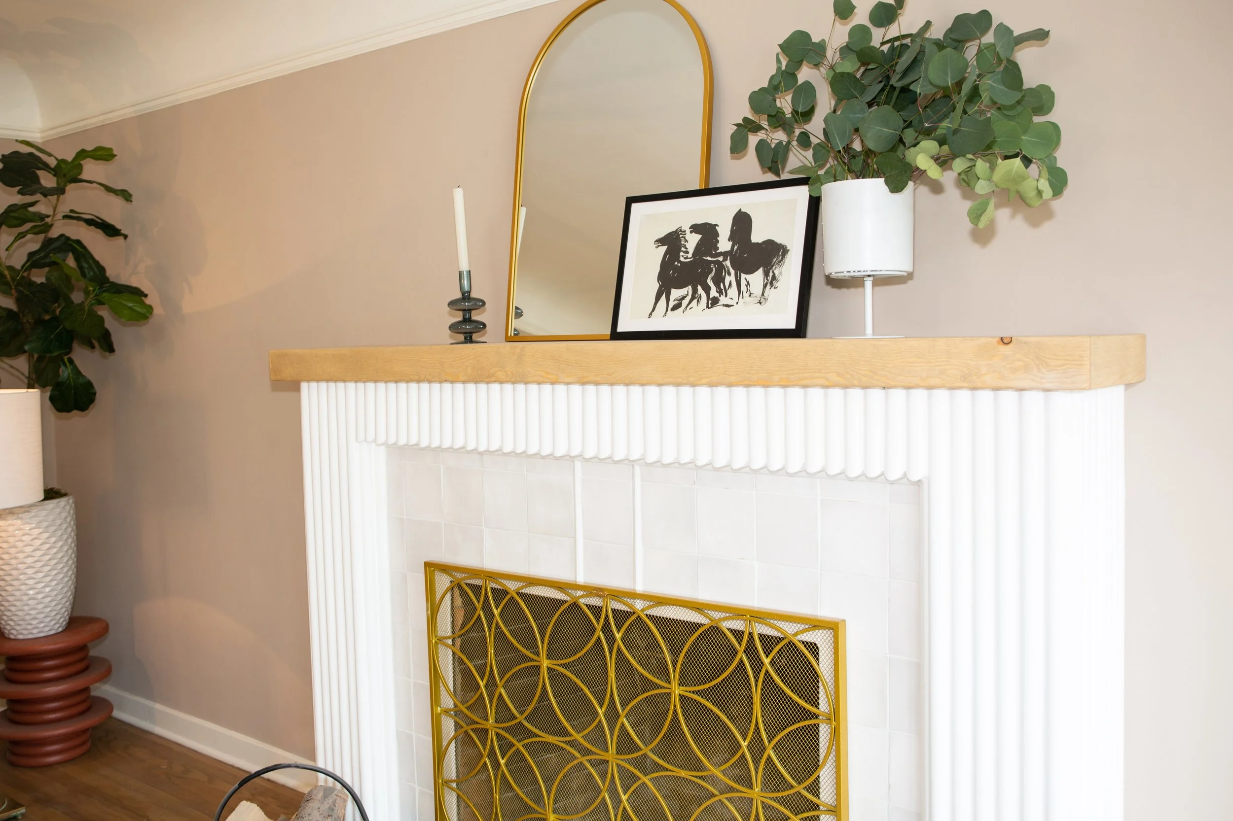 Living room fireplace mantel decorated with a gold mirror, black and white framed artwork of horses, a white potted plant, a candle holder with a white candle, and a black vase. Two large green plants are nearby.