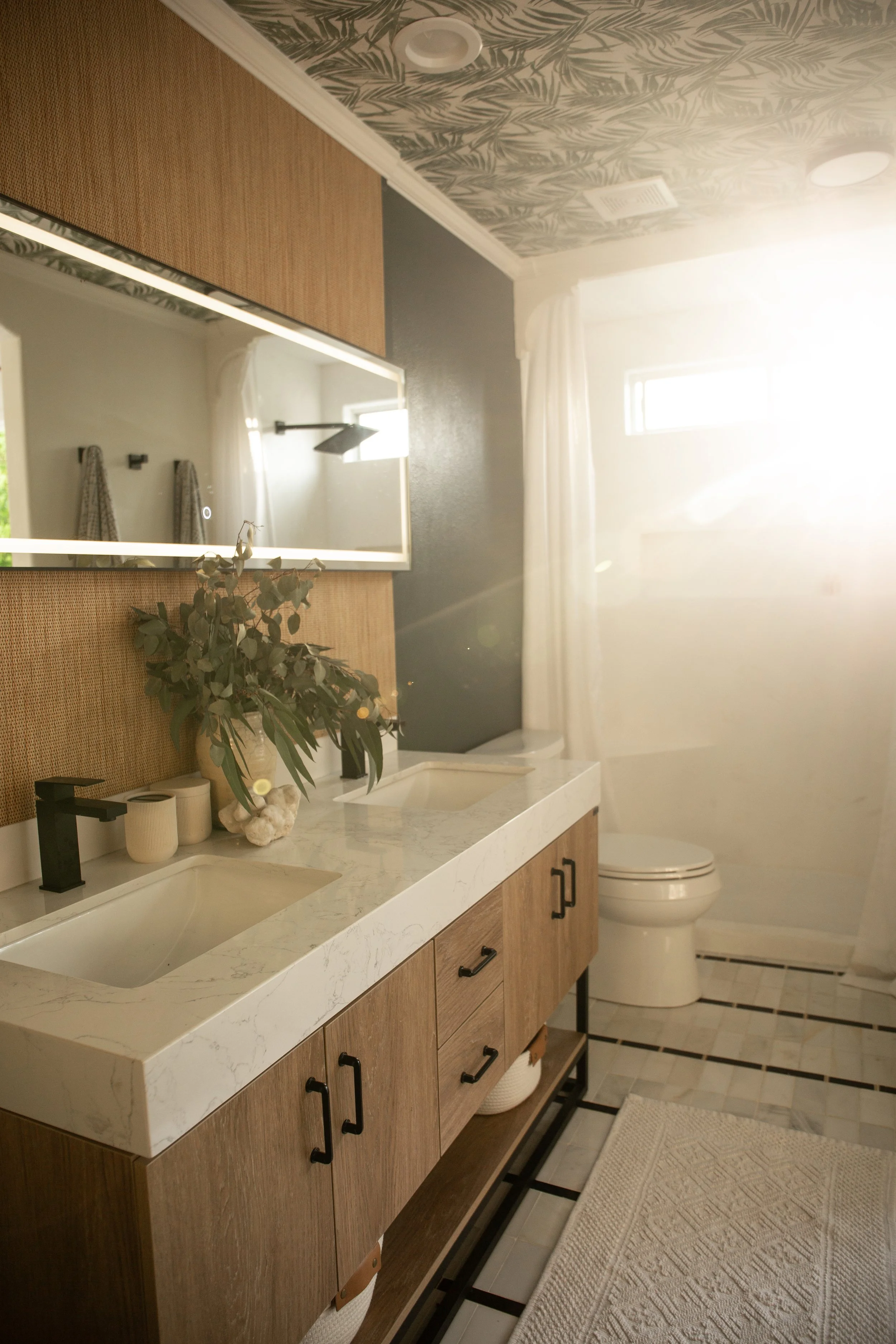 A modern bathroom with a dual sink vanity, large mirror, decorative plant, and a toilet, illuminated by natural sunlight from a window.