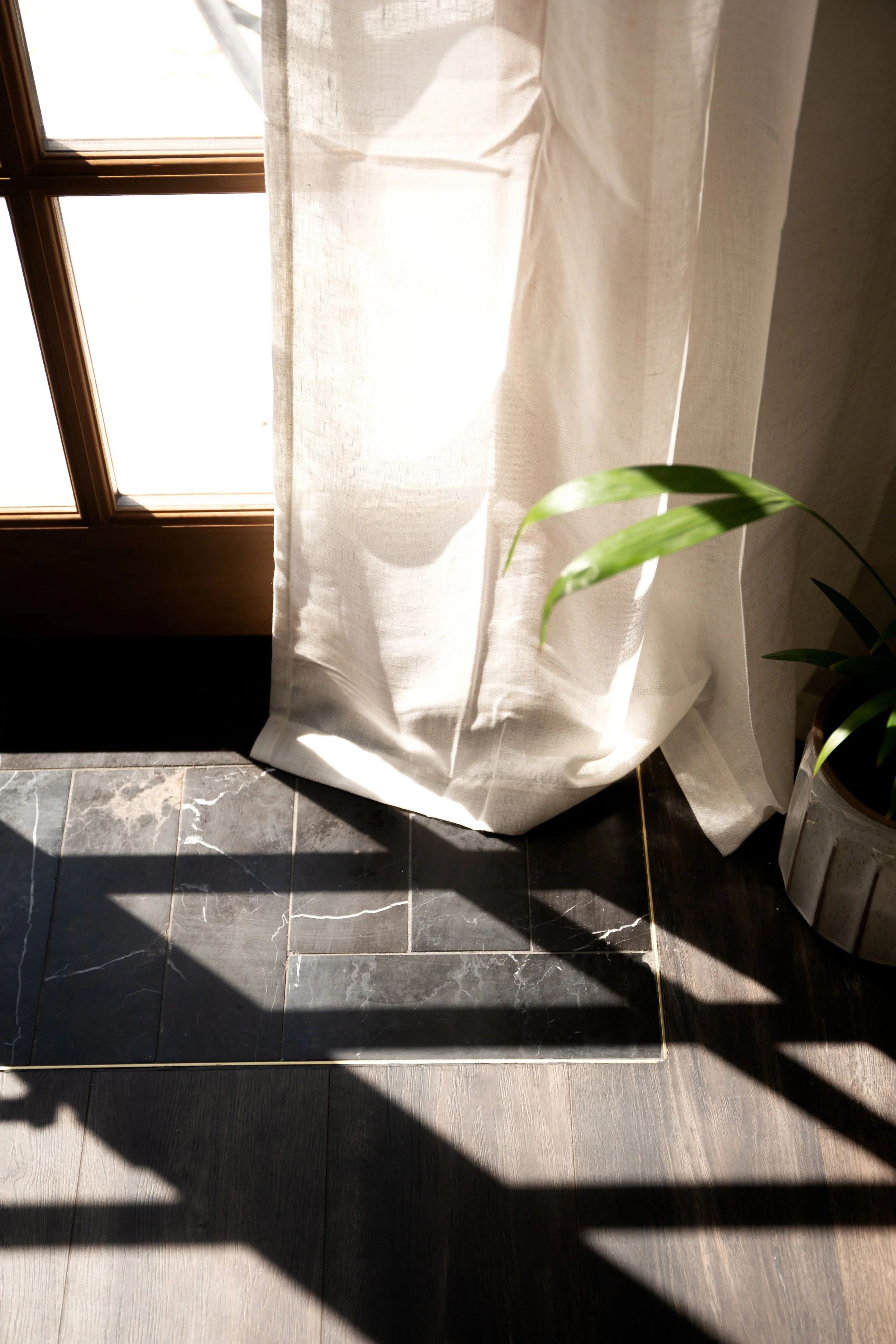 Sunlight streaming through a window with a sheer curtain, casting shadows on a tiled floor near a potted plant.