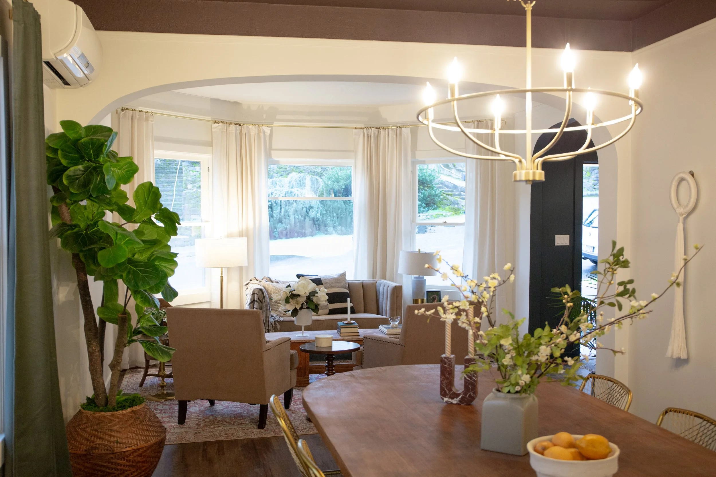 A cozy living room with large bay windows, white curtains, beige armchairs, and a beige sofa, decorated with flowers, lamps, and books. A large green potted plant is on the left, and a chandelier hangs above a wooden dining table with a ceramic vase 
