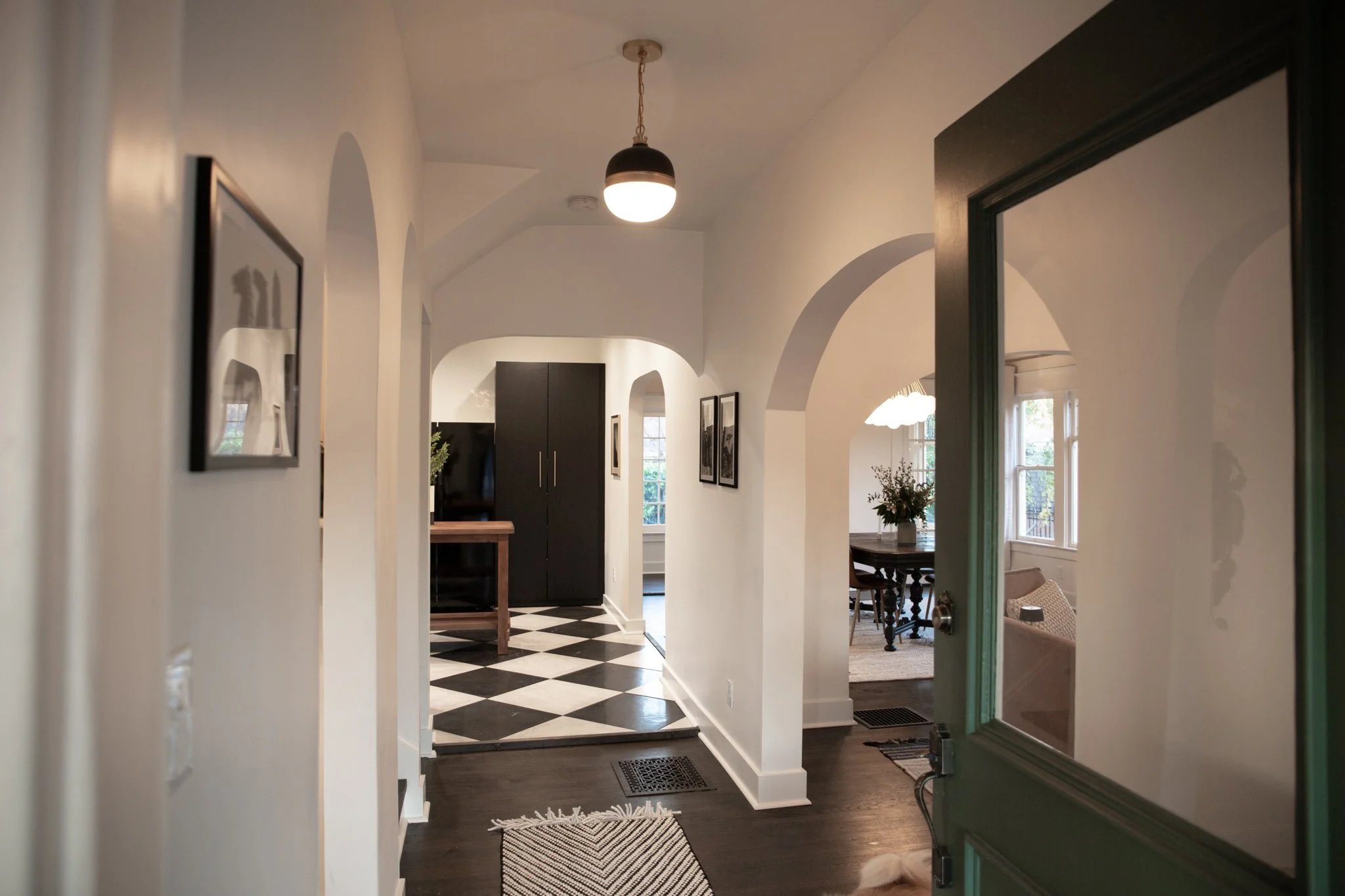 View of a homely hallway leading to a dining area with a dark cabinet, black and white checkered floor, and a table with a flower arrangement. The hallway has white walls, arched doorways, and a hanging ceiling lamp. The door in the foreground is gre