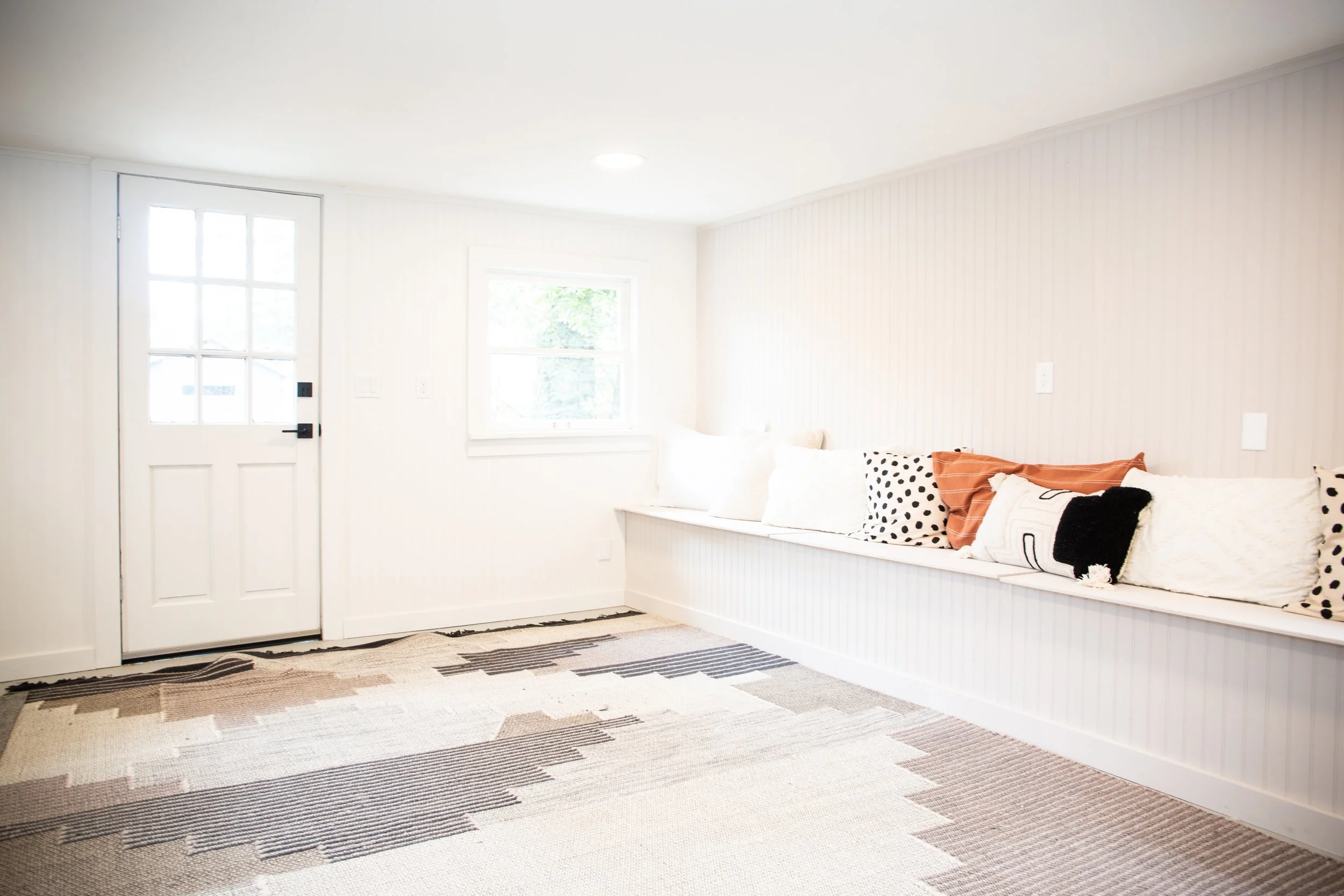 Bright entryway with a white door and a window, a built-in bench with decorative pillows, and a patterned rug on the floor.