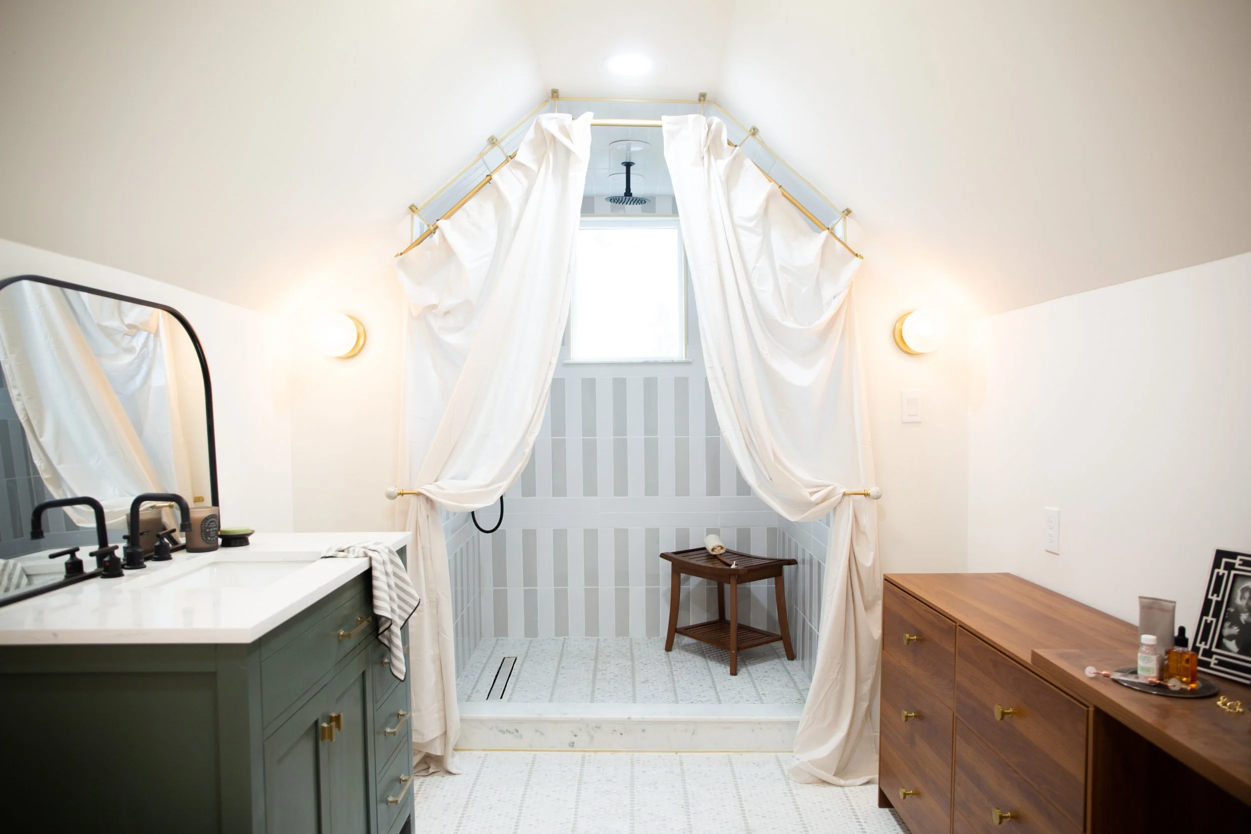 Bathroom with a walk-in shower area separated by white curtains, small window, vanity with a mirror and black fixtures, dresser with personal items, and a wooden stool inside the shower.