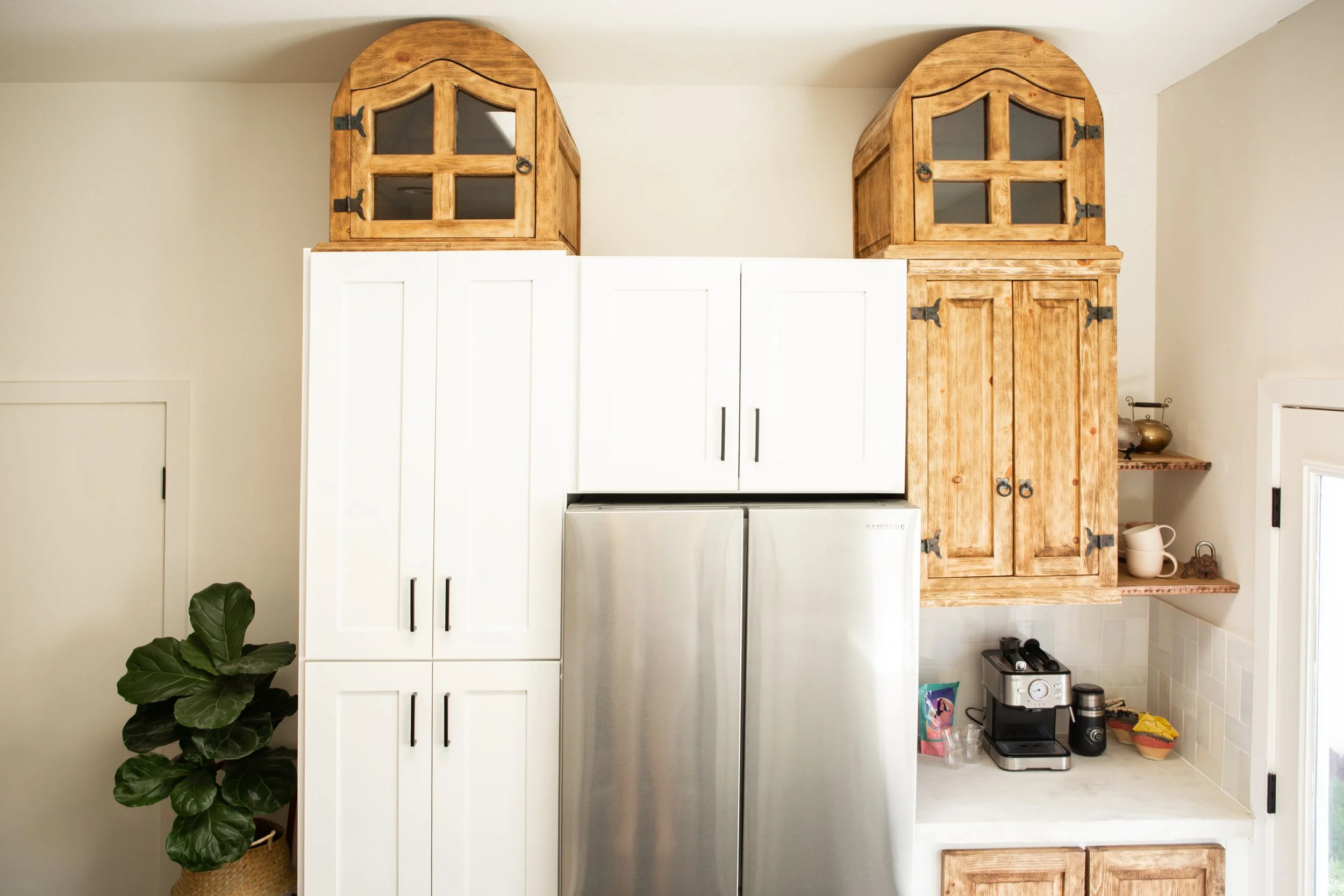 Kitchen upper cabinets with a stainless steel refrigerator below and wooden cabinets on top, featuring decorative black hinges and hardware. Open shelving with small kitchen items, including a coffee maker and cups, next to a window.