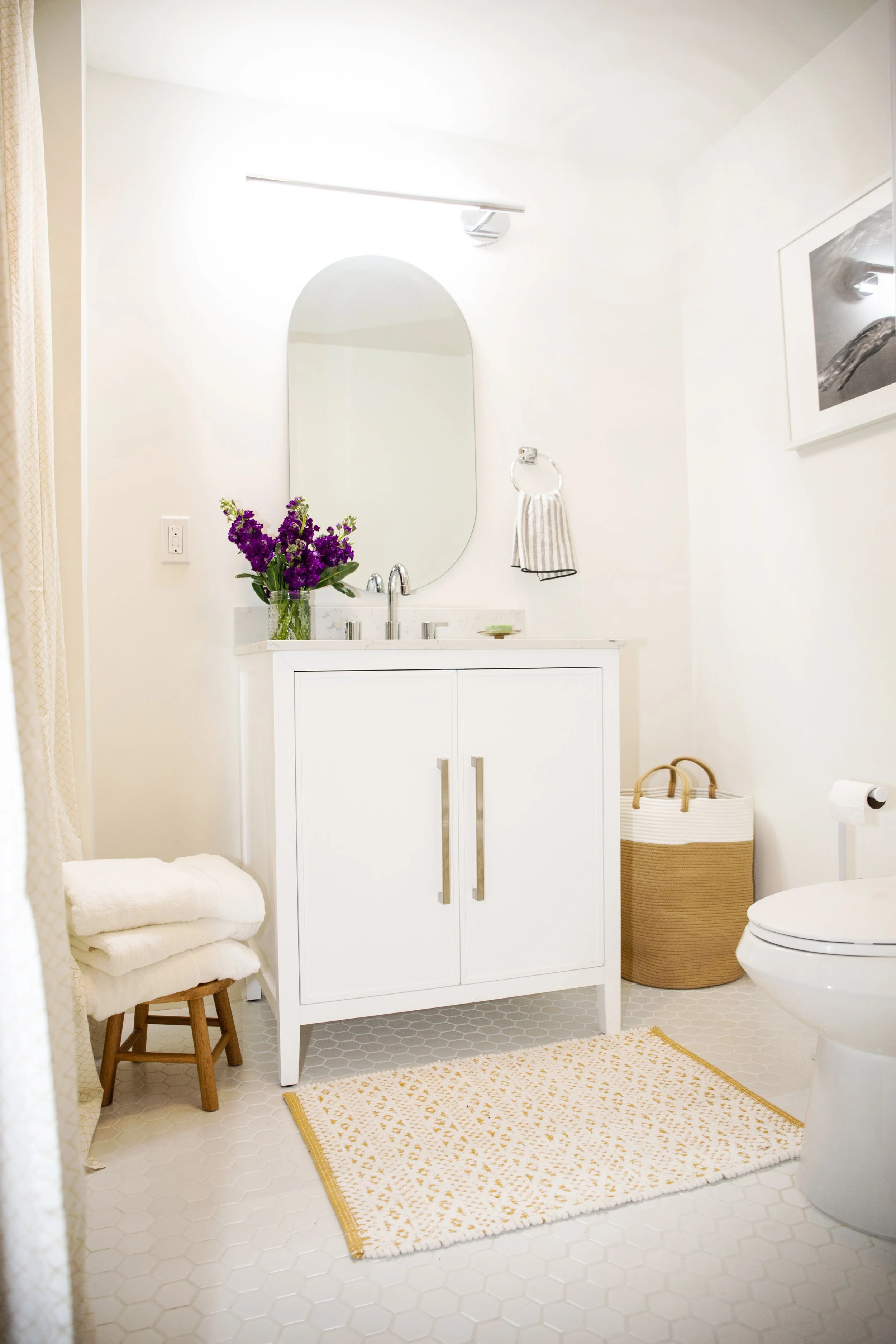 A bright, minimalist bathroom with a white vanity, mirror, and hexagonal tile flooring, decorated with a vase of purple flowers, a basket, towels, and wall art.