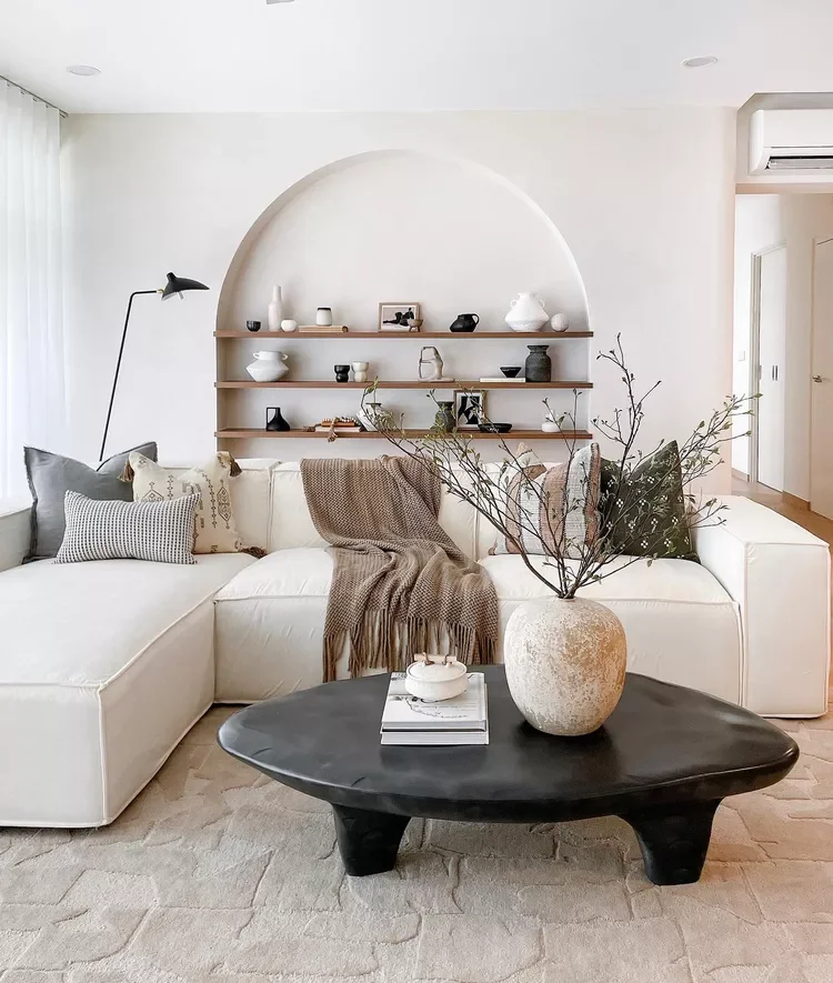 Living room with white sectional sofa adorned with patterned pillows and a brown blanket, a black coffee table with a large vase and branch arrangement, a wall with built-in shelves holding decorative items, and a floor lamp.