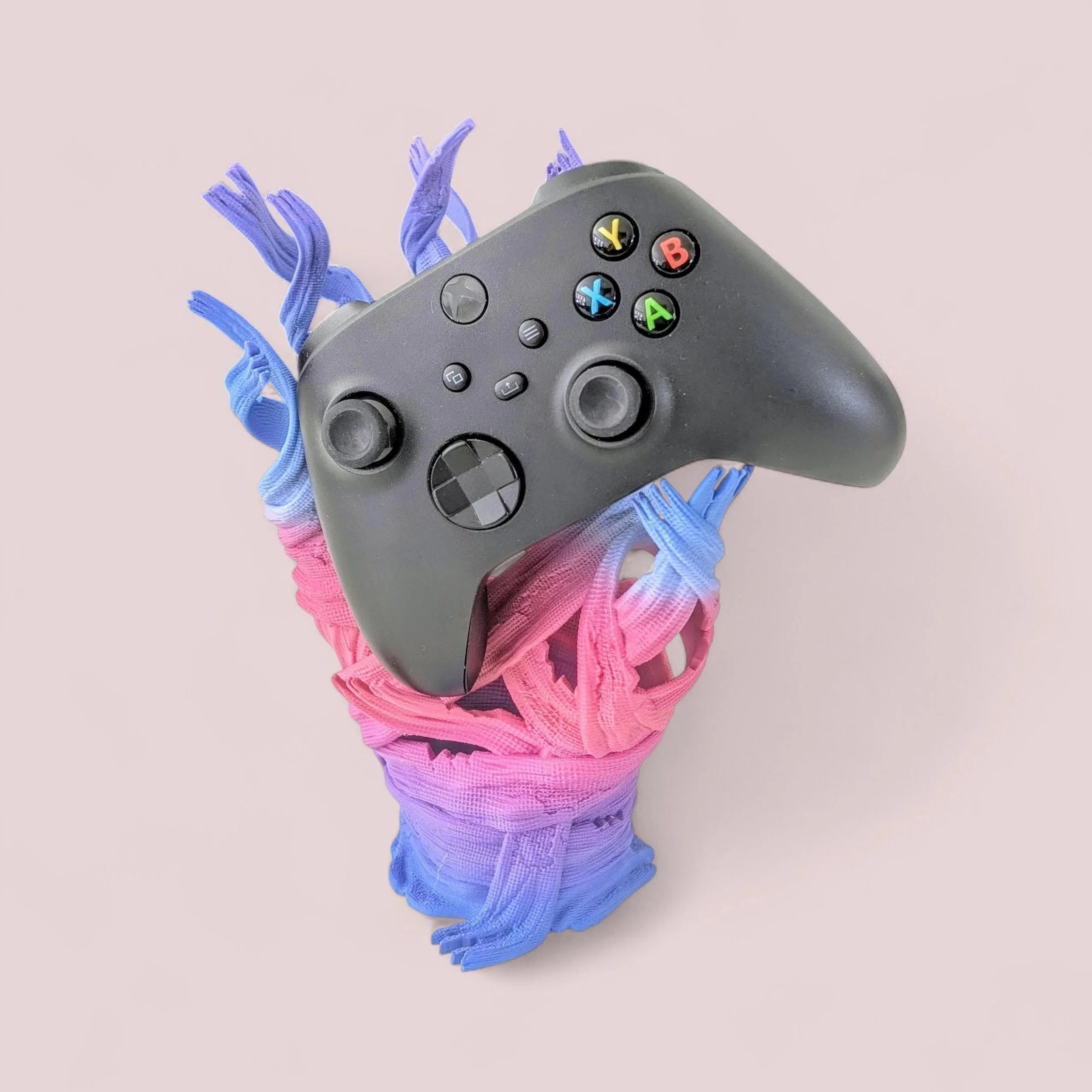 A video game controller resting on a multicolored mummy hand controller stand, with pink, purple, and blue sections, on a light pink background.