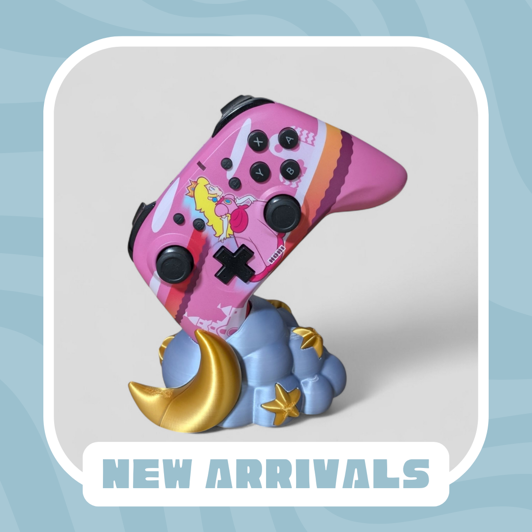 A gaming controller with a pink design featuring peach, mounted on a 3D printed decorative cloud with a golden crescent moon and stars, with the text "New Arrivals" at the bottom.