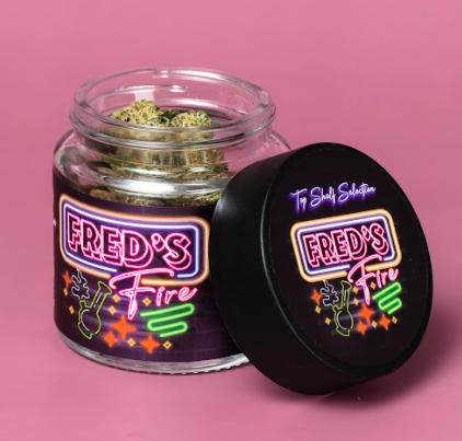 Open jar of cannabis with colorful label reading "FRED'S Fire" against a pink background.