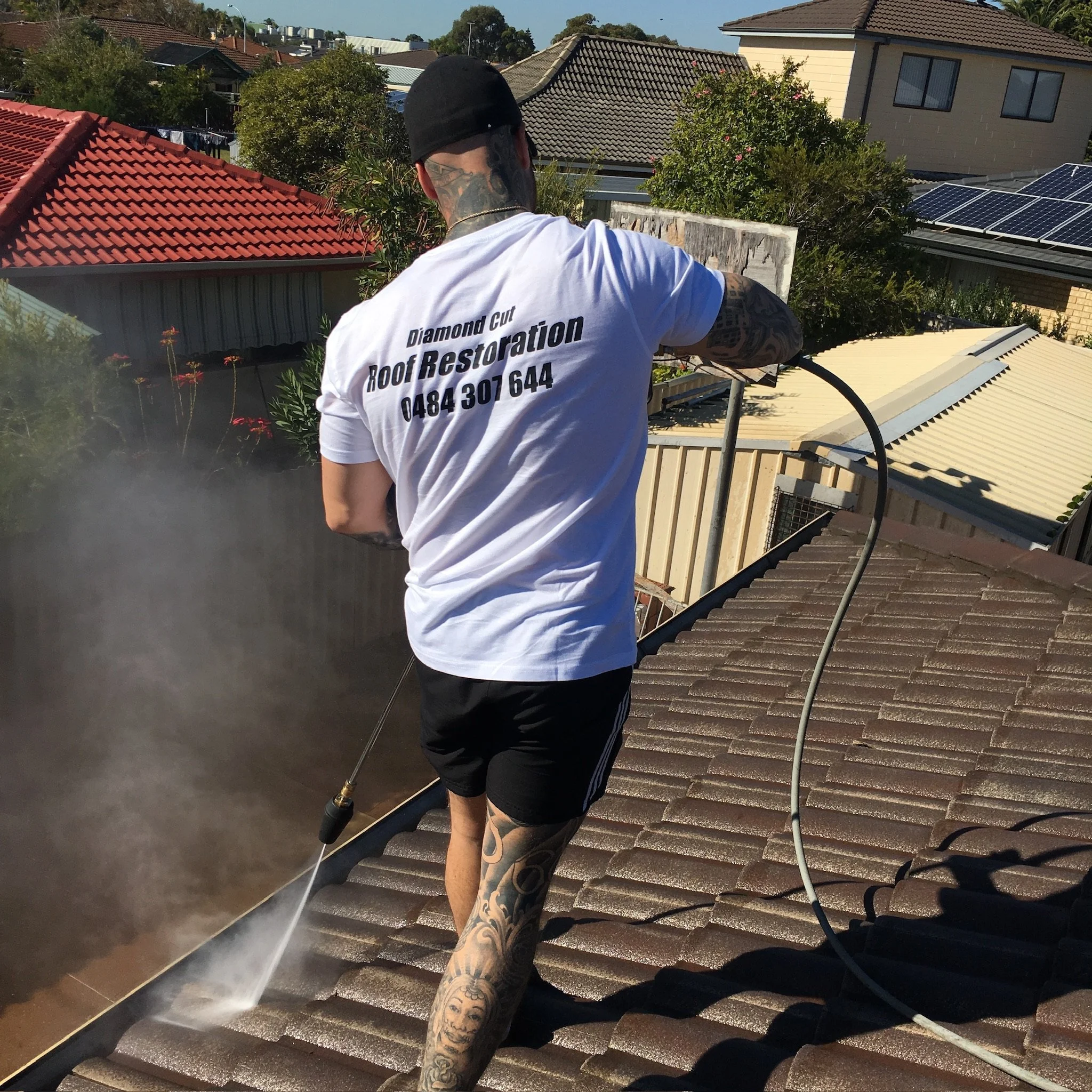 A man with tattoos on his arms and legs, wearing a black cap and a white T-shirt that says 'Diamond Cut Roof Restoration' with a phone number, is pressure washing a tiled roof on a sunny day. The roof has brown tiles, and the background shows neighboring houses, trees, and solar panels.