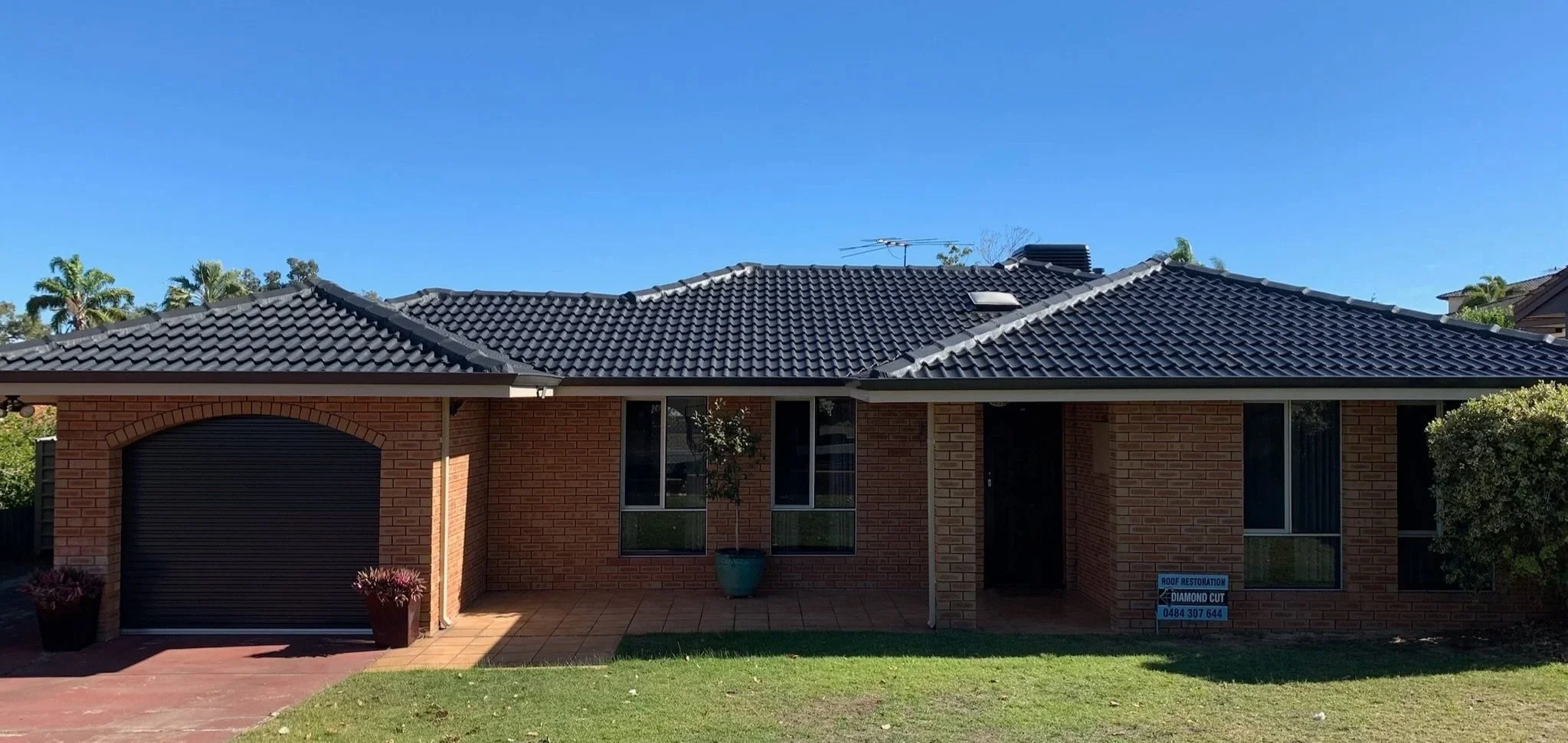 A brick house with a black tiled roof, two front windows, and a closed garage door. There are potted plants near the front of the house and a sign advertising roof restoration services.