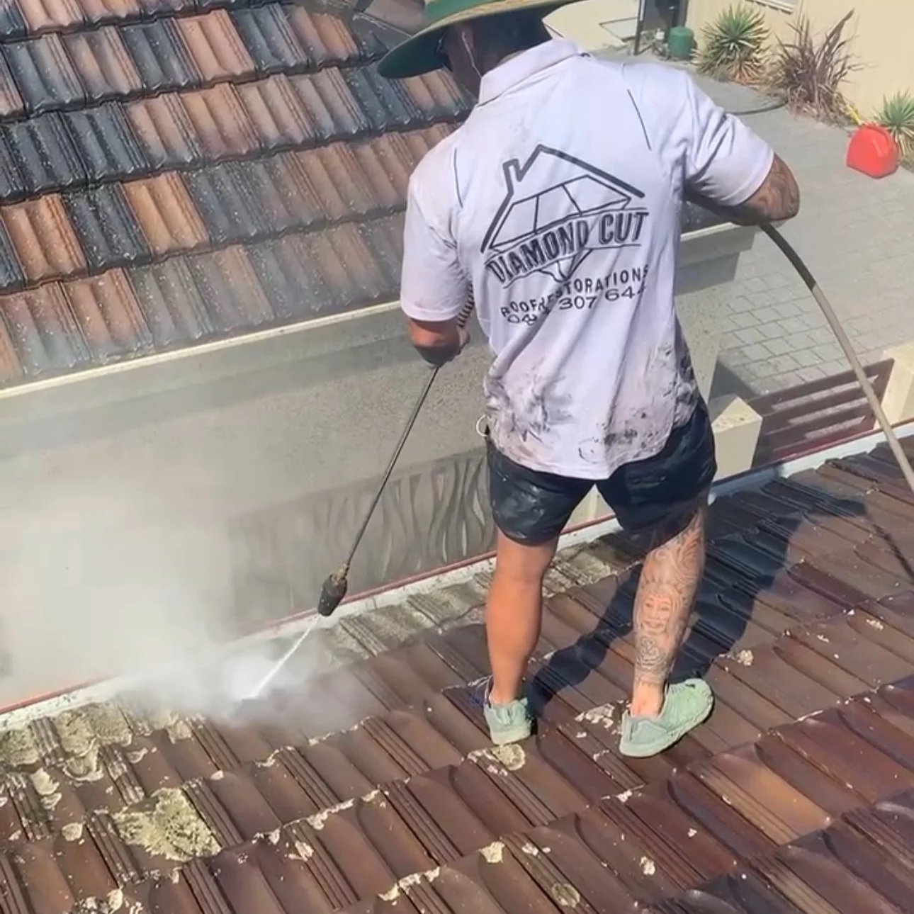 A worker in a diamond cut roofing company shirt pressure washing a tiled roof. The worker has tattoos on one leg and is wearing a hat, green shoes, and black shorts.