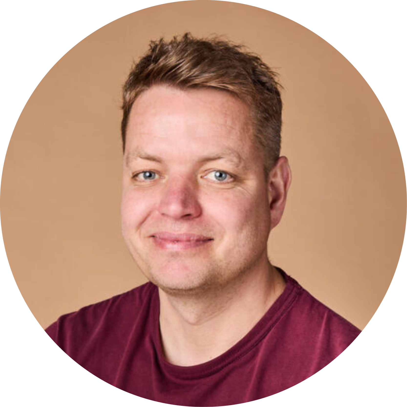 Portrait of a young man with short brown hair, light skin, blue eyes, smiling slightly, wearing a maroon shirt, against a beige background.