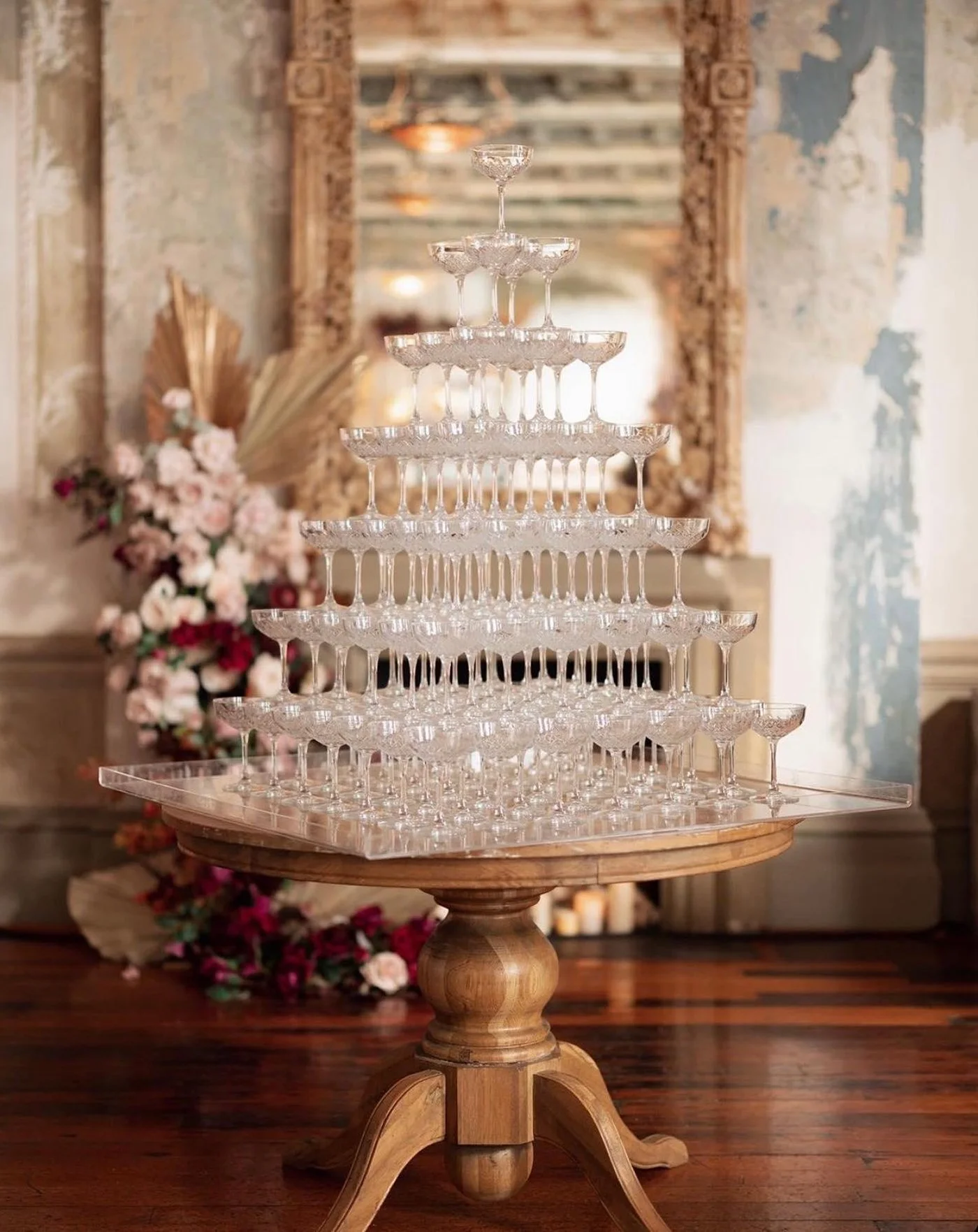 A pyramid of stacked champagne glasses on a wooden table with a floral arrangement and mirror in the background.