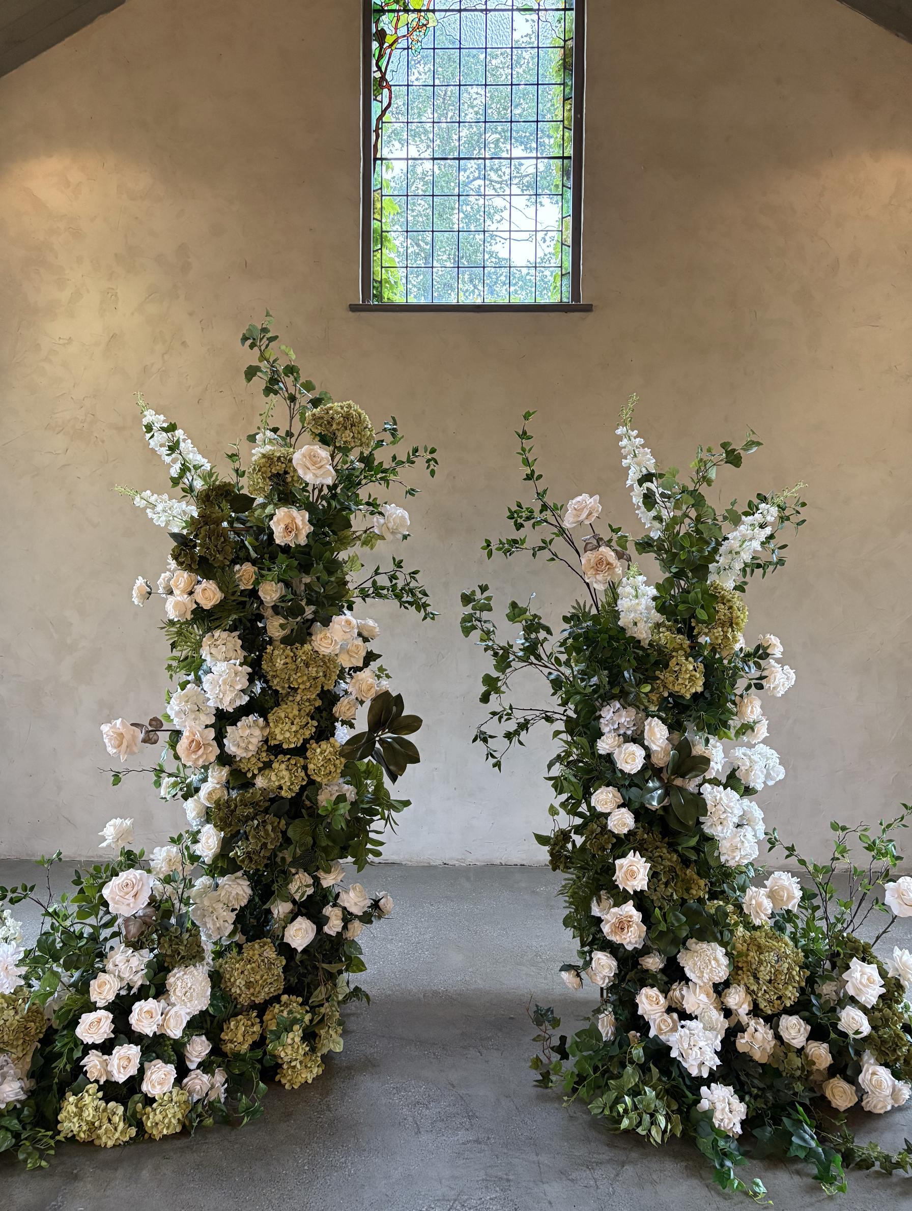 Floral arrangement with white roses, hydrangeas, and greenery inside a building with a stained glass window.