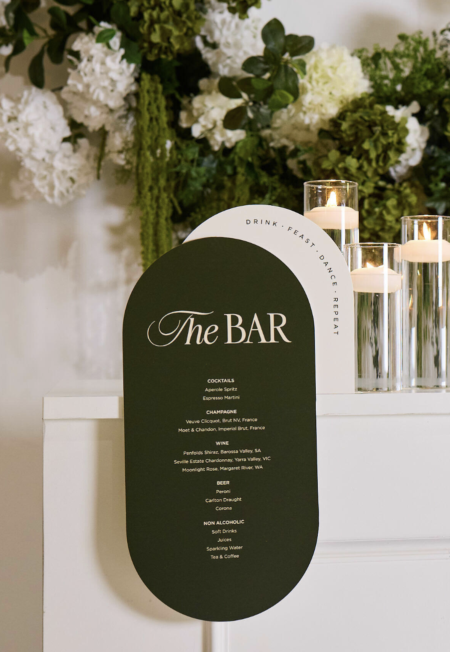 A black and white sign with the heading 'The BAR' listing cocktails, champagne, wine, beer, and non-alcoholic drinks, set in front of a floral arrangement with candles.