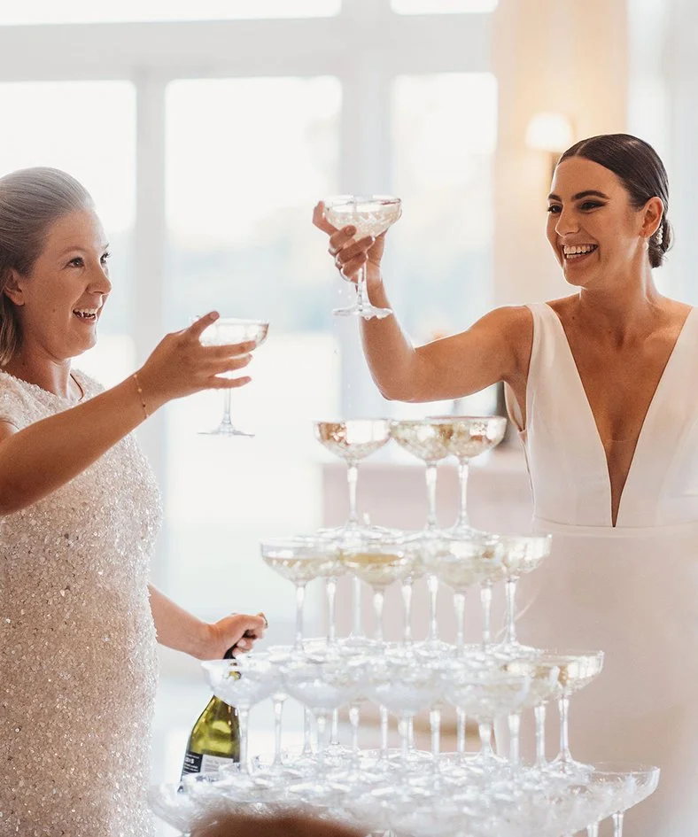 Two women celebrating with champagne glasses at a wedding or formal event, pouring drinks into a champagne tower pyramid.